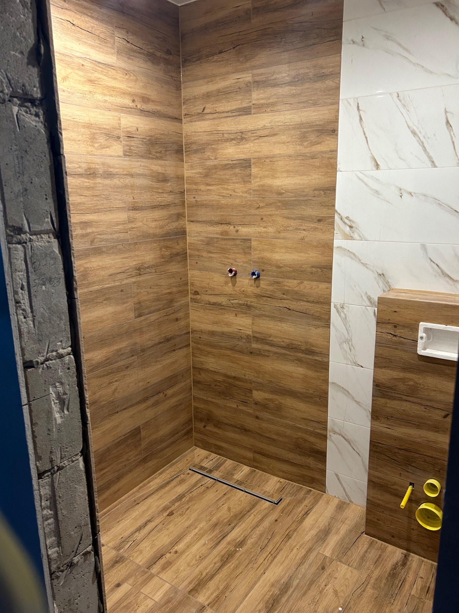 Prysznic typu walk-in z drewnopodobnymi płytkami na ścianach i podłodze, widoczny odpływ liniowy, fragment surowej ściany z betonu komórkowego i biało-marmurowe płytki na ścianie w tle.