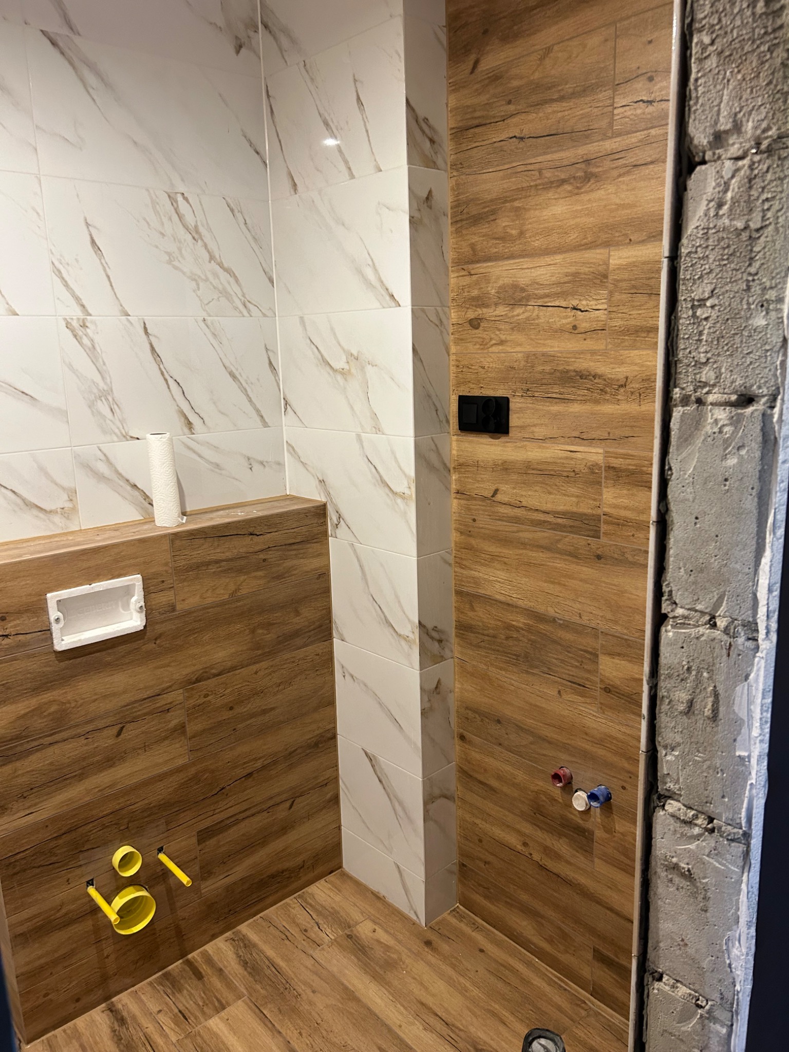 Wnętrze łazienki w trakcie remontu z częściowo wykończonymi ścianami: połączenie białych płytek z marmurkowym wzorem i drewnopodobnych paneli, widoczne instalacje wodno-kanalizacyjne i surowa...