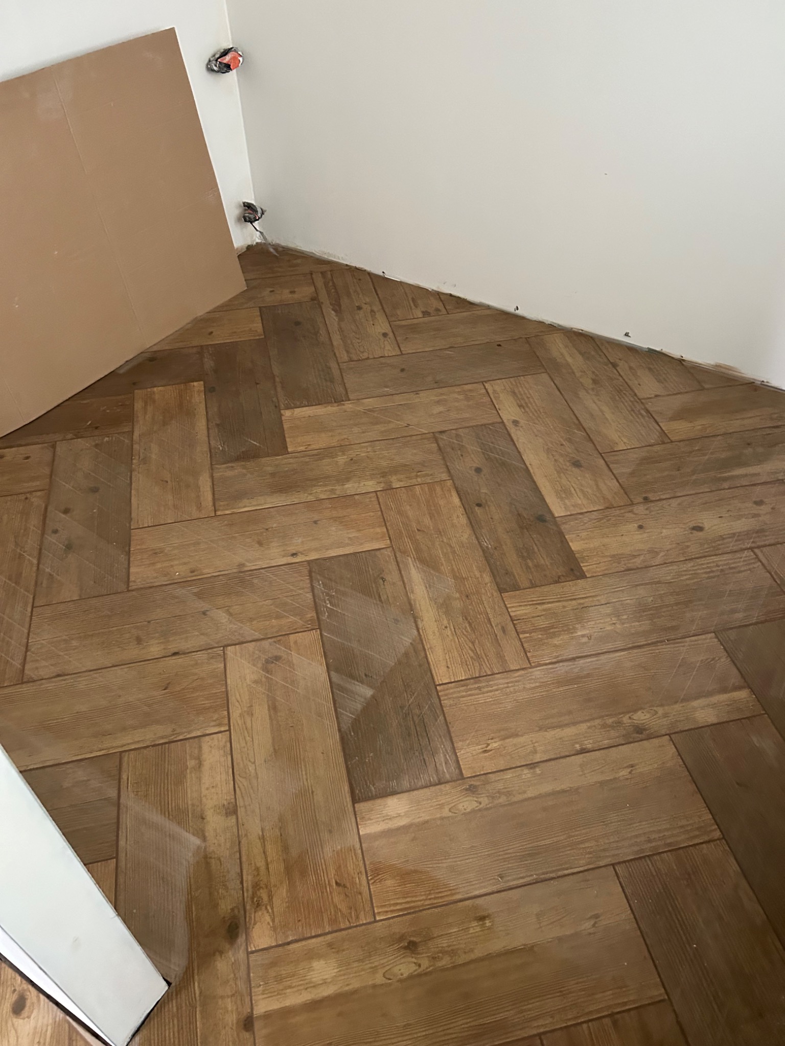 Ułożona podłoga z paneli imitujących drewno w jodełkę, widoczne niedokończone prace przy ścianie i wystające kable elektryczne.