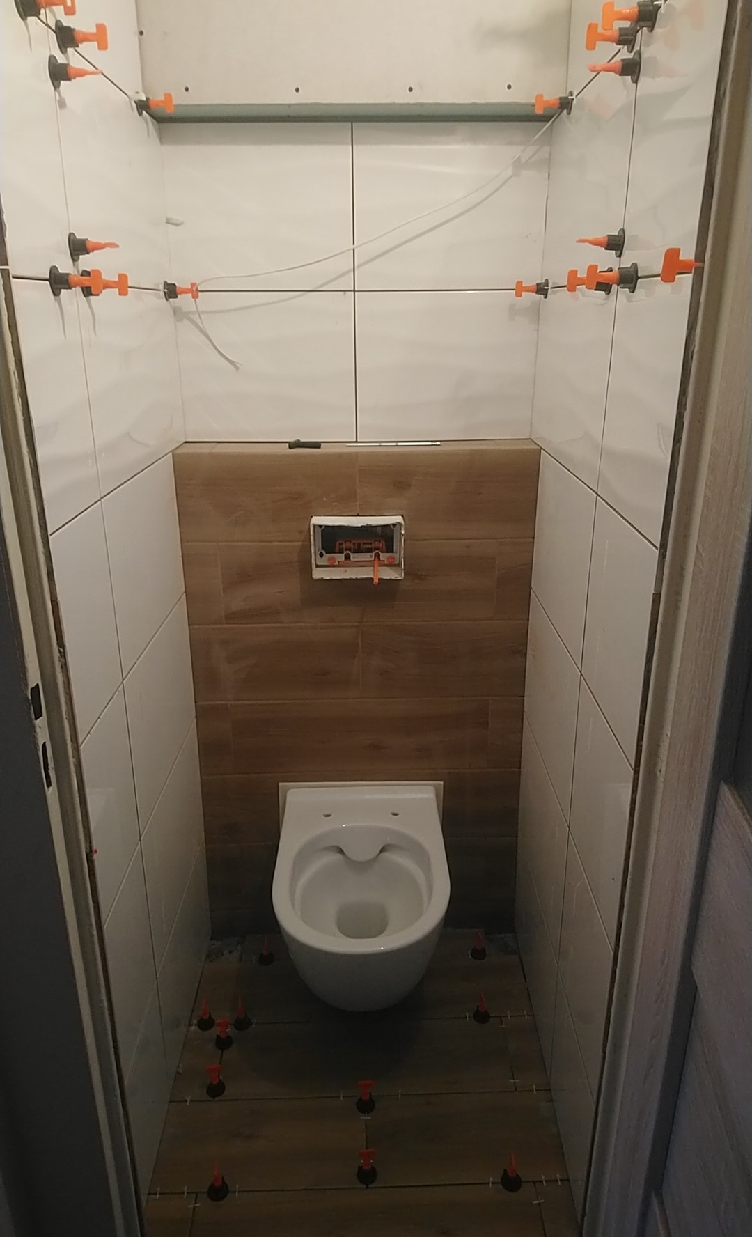 Montaż płytek w małej łazience z użyciem systemu poziomowania, widoczna toaleta i częściowo wykończona ściana z płytek imitujących drewno.