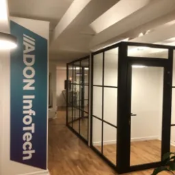 Biuro z przeszklonymi ścianami działowymi w czarnych ramach, widok na korytarz i logo firmy na filarze.