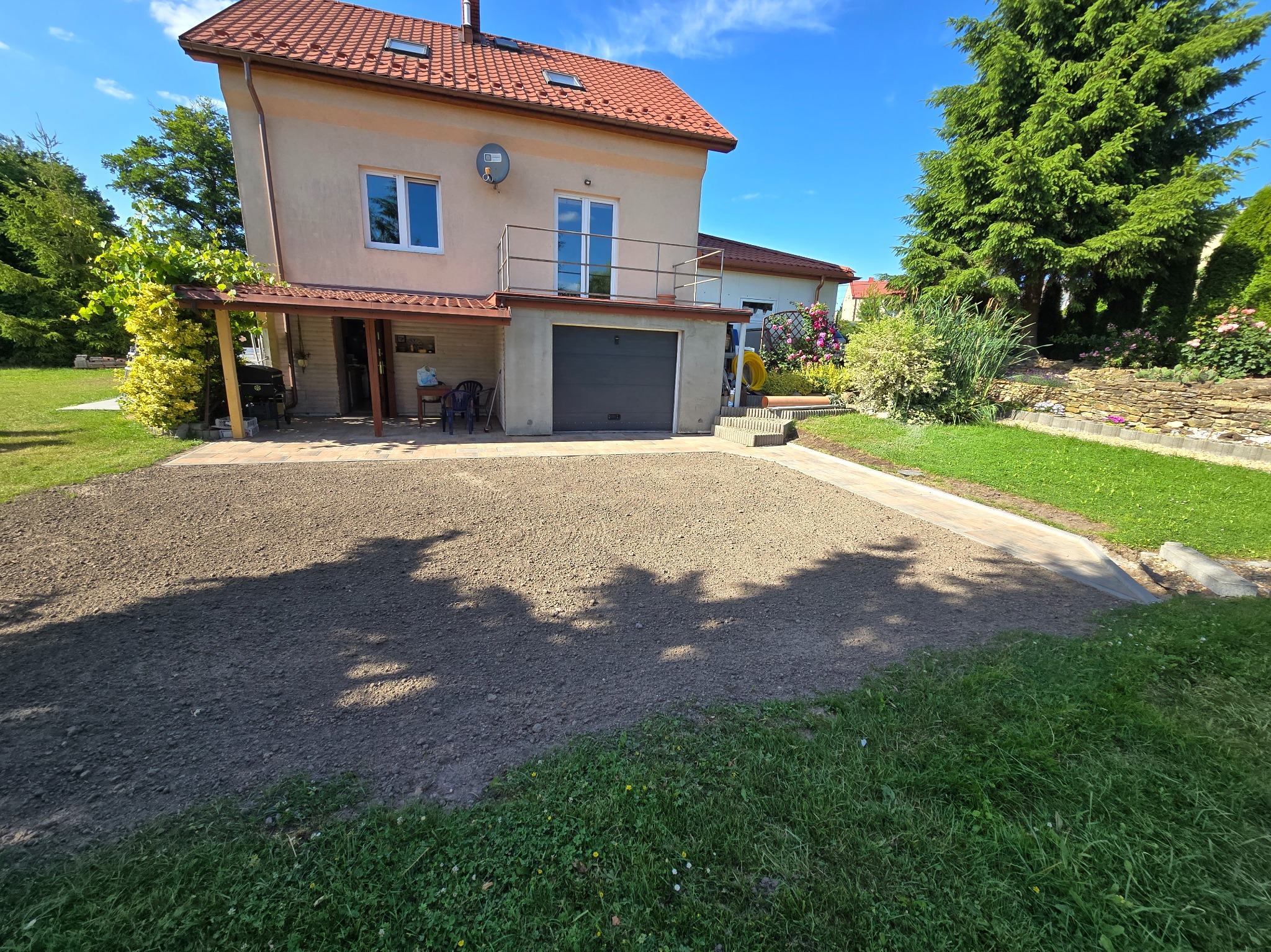 Przygotowany teren pod kostkę brukową przed domem z garażem. Widoczne wyrównane podłoże, fragment trawnika i elewacja budynku z balkonem.