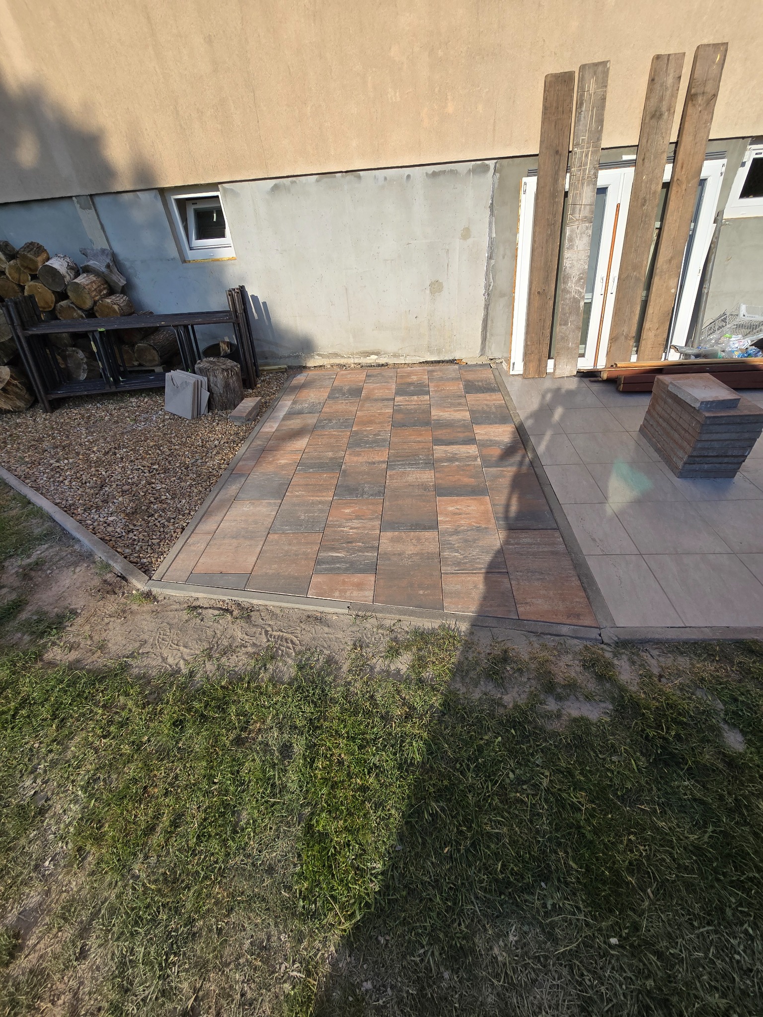 Ułożona kostka brukowa w odcieniach brązu i szarości, przylegająca do budynku. Część terenu wyłożona płytkami, obok stos kostki. Widoczne drewniane deski i trawa.