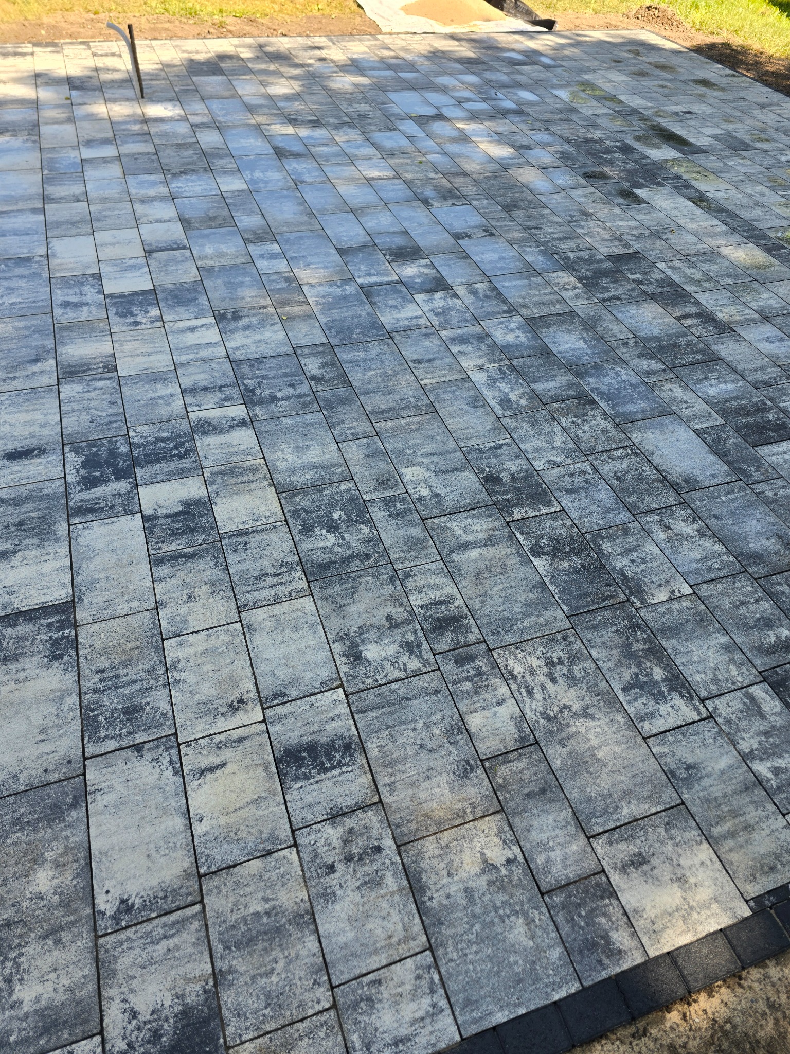 Szara kostka brukowa ułożona na dużej powierzchni z widocznym obrzeżem. Widok z góry ukazuje wzór ułożenia i fakturę kostki. W tle trawnik.