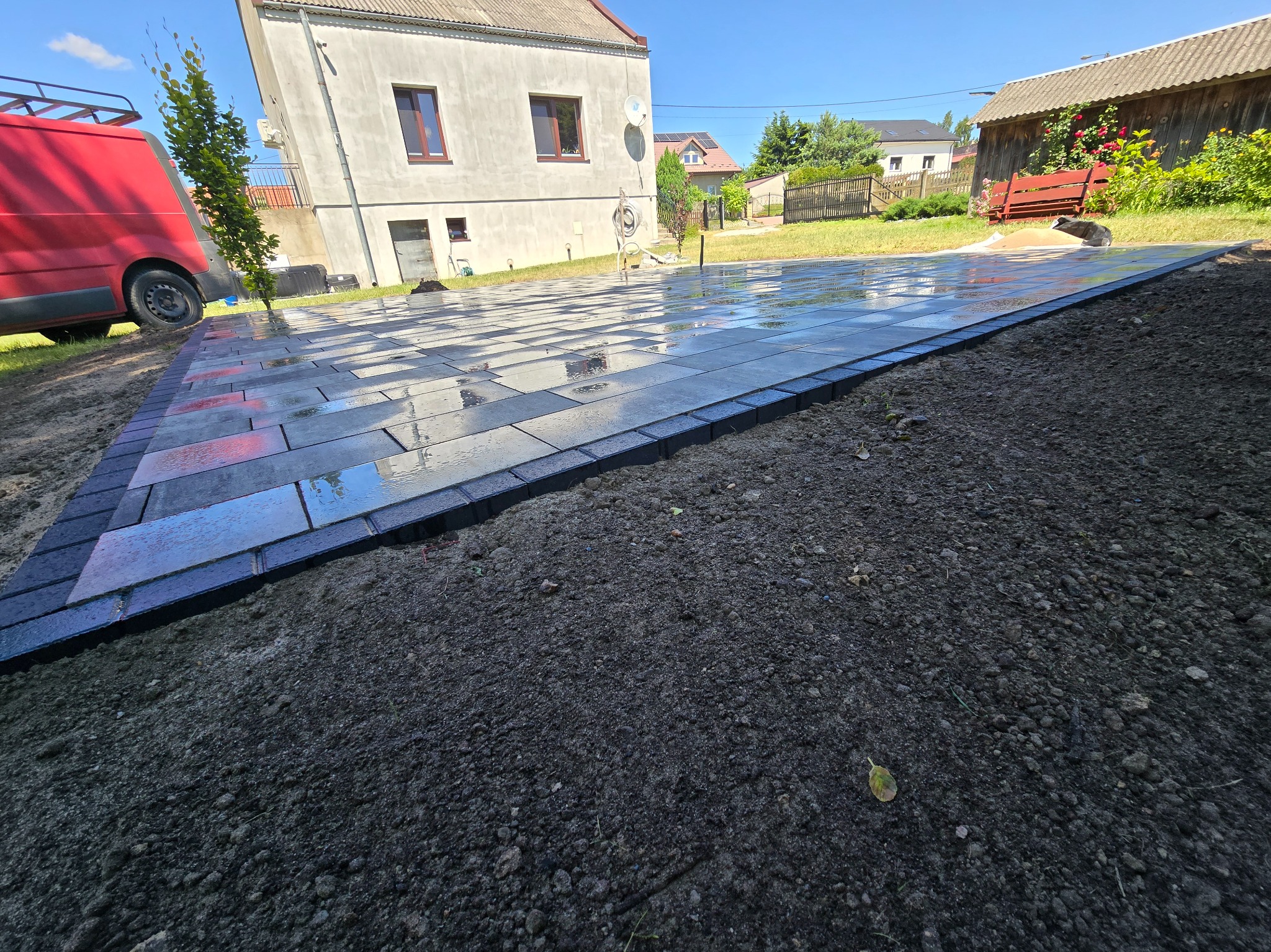 Świeżo ułożona, mokra kostka brukowa w odcieniach szarości, obramowana ciemniejszymi kostkami. W tle budynki mieszkalne i częściowo widoczny czerwony samochód.