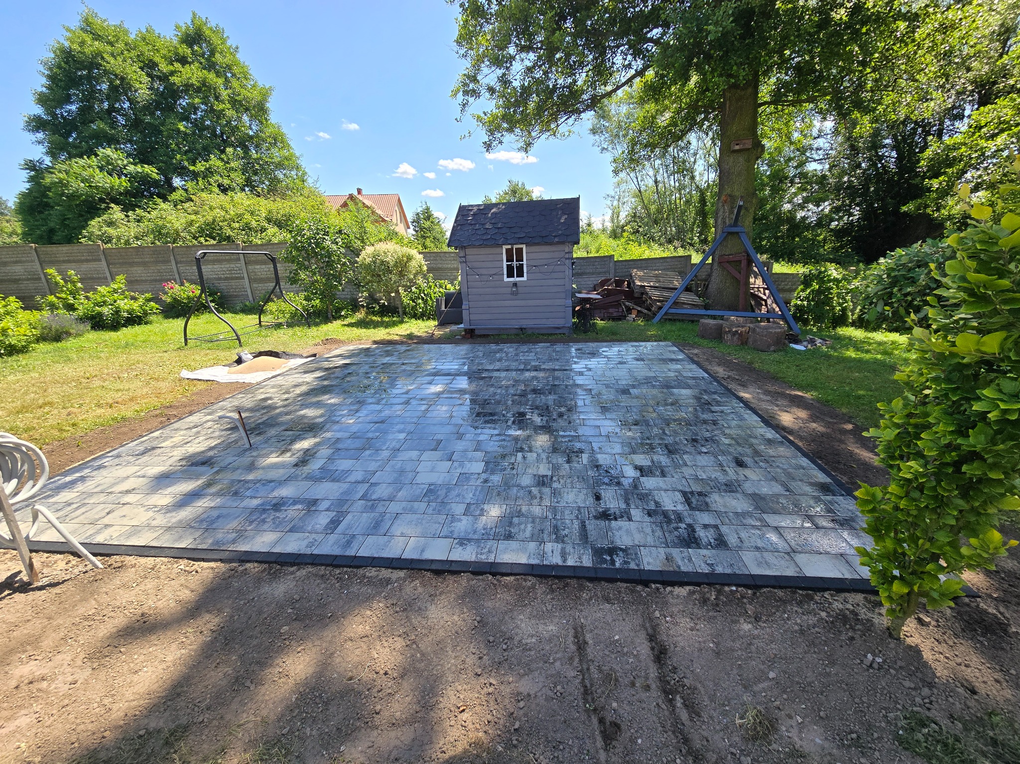 Ułożona, prostokątna nawierzchnia z szarej kostki brukowej w ogrodzie, obok małego domku narzędziowego i huśtawki. Widoczne obrzeża i przygotowane podłoże.