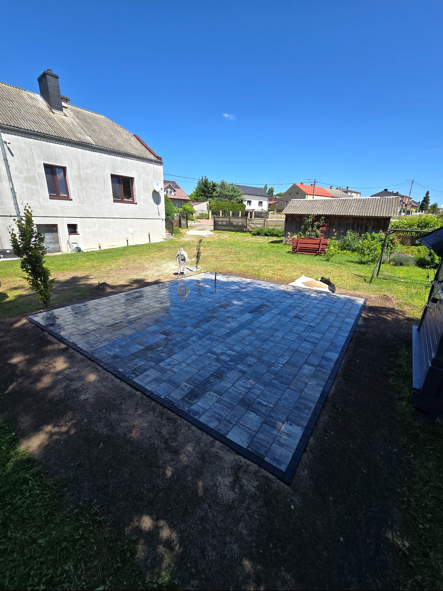 Nowa, kwadratowa nawierzchnia z szarej kostki brukowej z czarnym obramowaniem na trawniku, dom w tle. Widok z góry, słoneczny dzień.