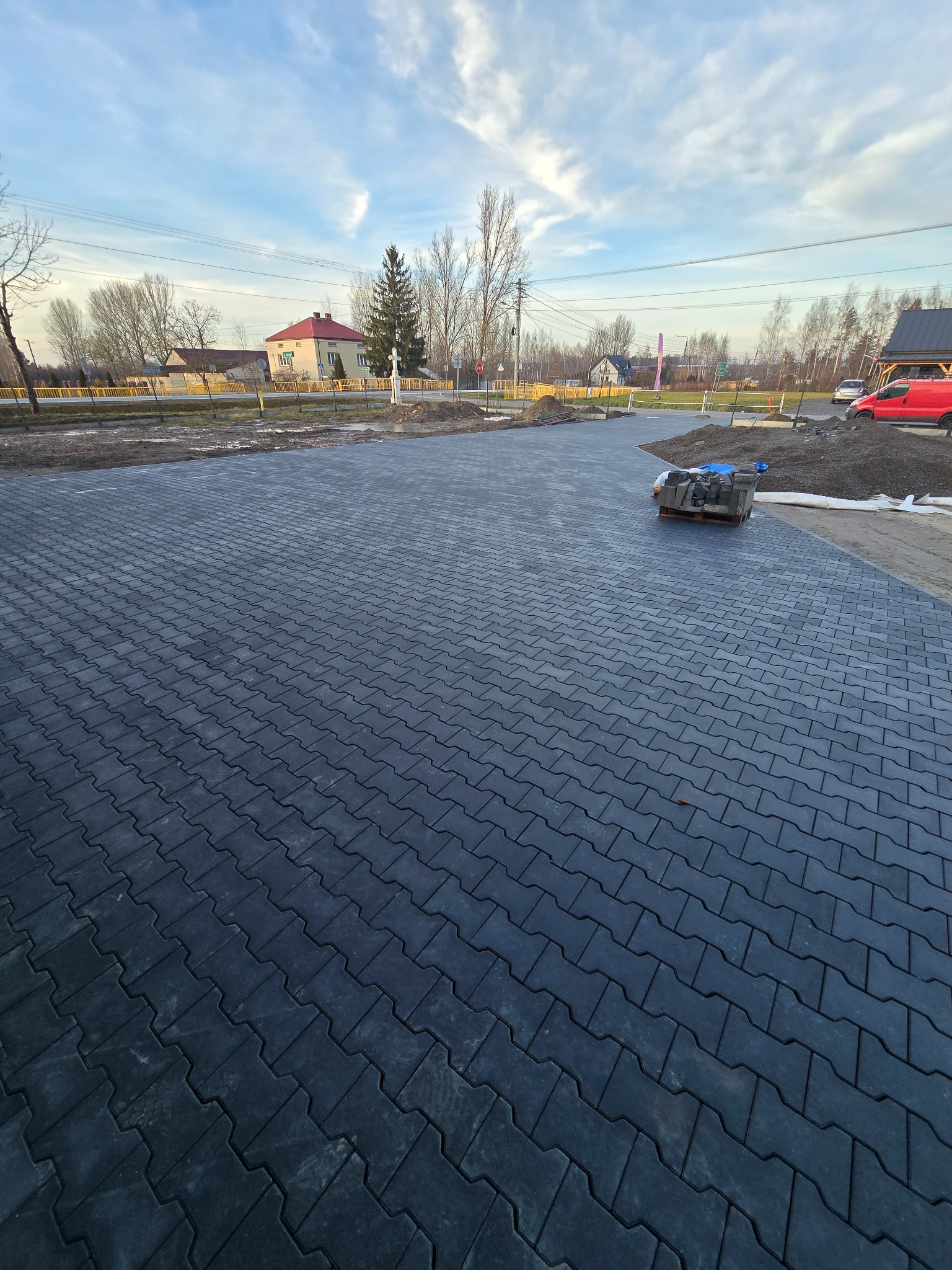 Nowo ułożona, ciemnoszara kostka brukowa na dużej powierzchni, z paletą kostki w tle. Widoczne domy i drzewa w oddali, błękitne niebo z chmurami.