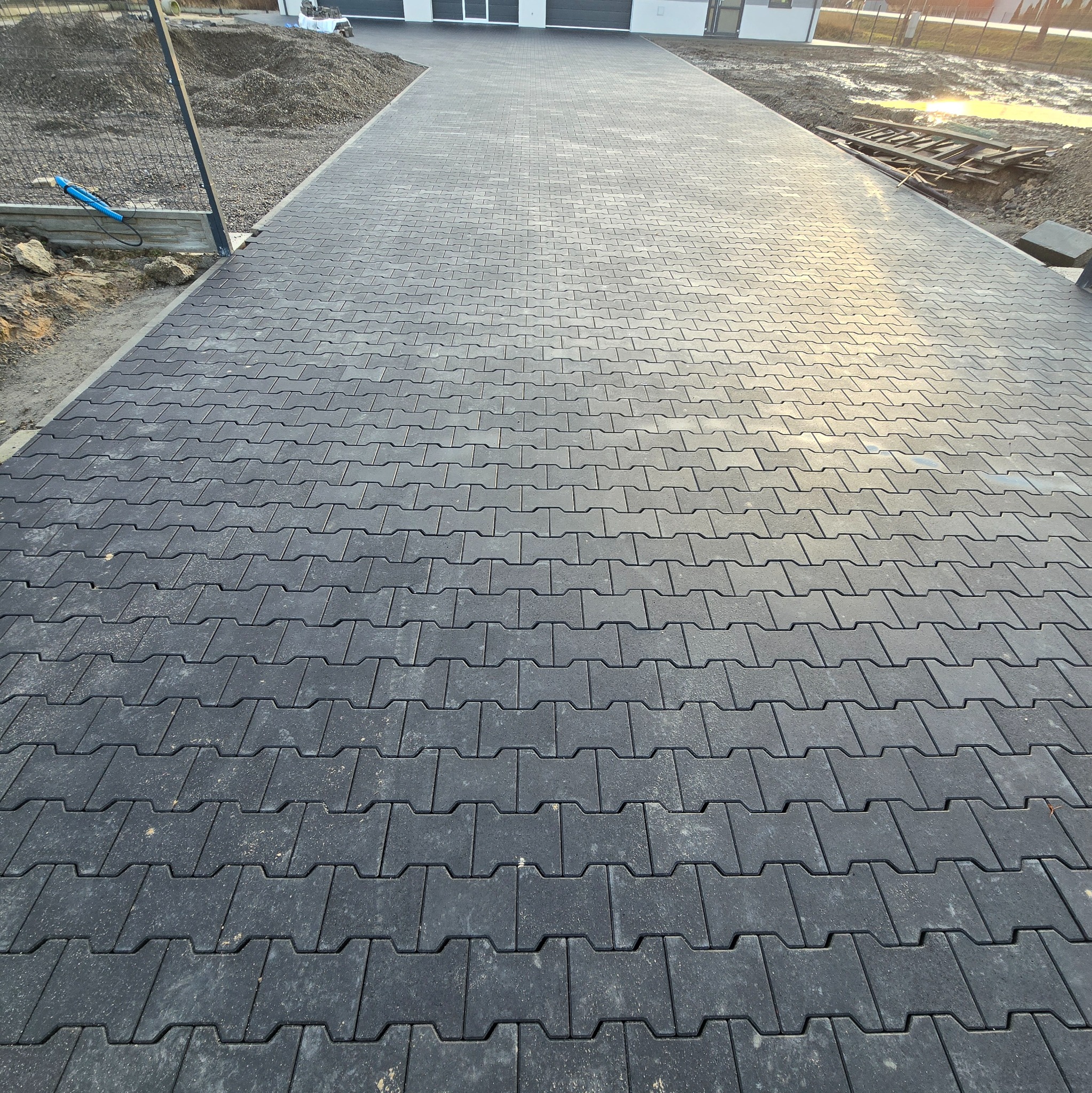 Ułożona, szara kostka brukowa typu I na podjeździe do domu. Widoczne przygotowane podłoże i materiały budowlane. Słoneczne oświetlenie podkreśla strukturę kostki.