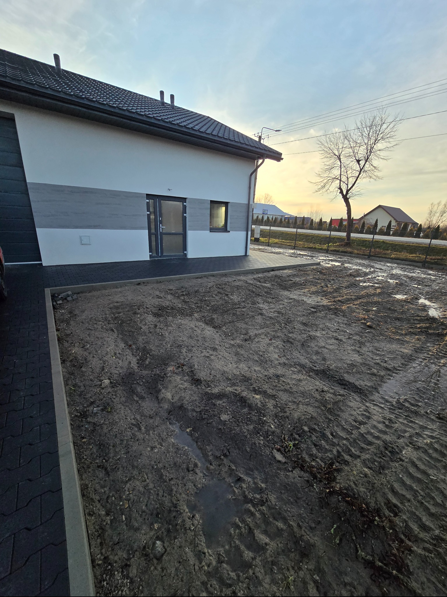 Nowoczesny dom z szarą kostką brukową i przygotowanym terenem pod trawnik. Widoczne ślady opon i kałuże na ziemi. W tle domy jednorodzinne i drzewo bez liści.
