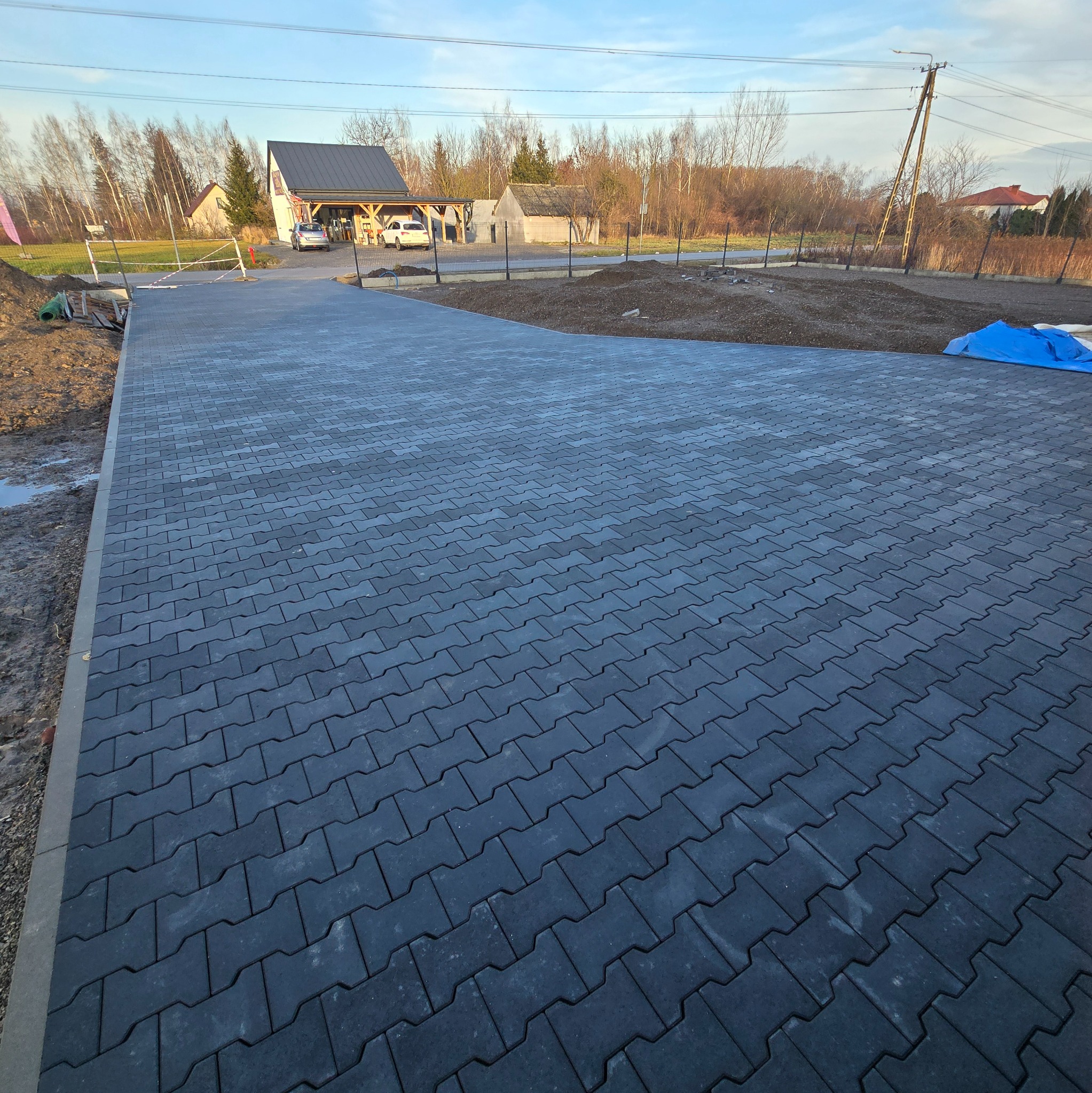 Nowa, ciemnoszara kostka brukowa ułożona na posesji. Widoczne budynki w tle i przygotowany teren pod dalsze prace. Dobre oświetlenie, szeroki kadr.