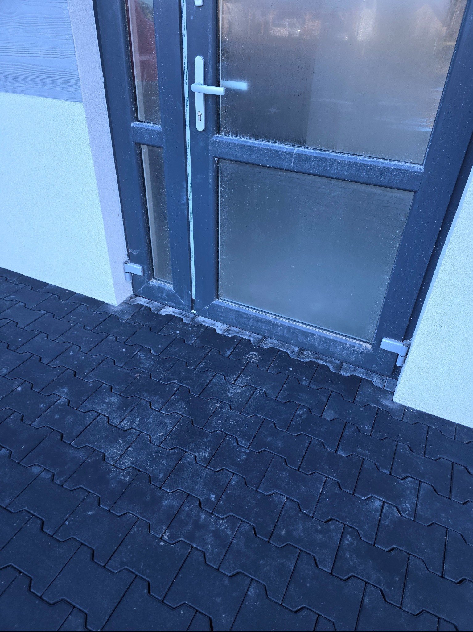 Ciemna kostka brukowa ułożona przy szarych drzwiach wejściowych. Widoczne detale łączeń i struktura kostki. Minimalistyczny styl.