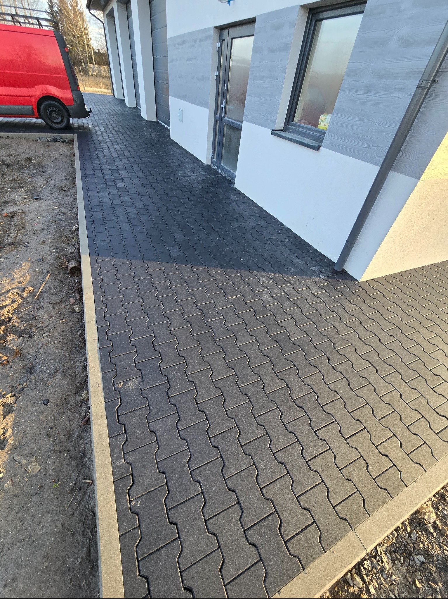 Ułożona, grafitowa kostka brukowa przed budynkiem z szarą elewacją. Widoczny fragment czerwonego samochodu dostawczego. Krawężnik oddziela kostkę od surowego gruntu.