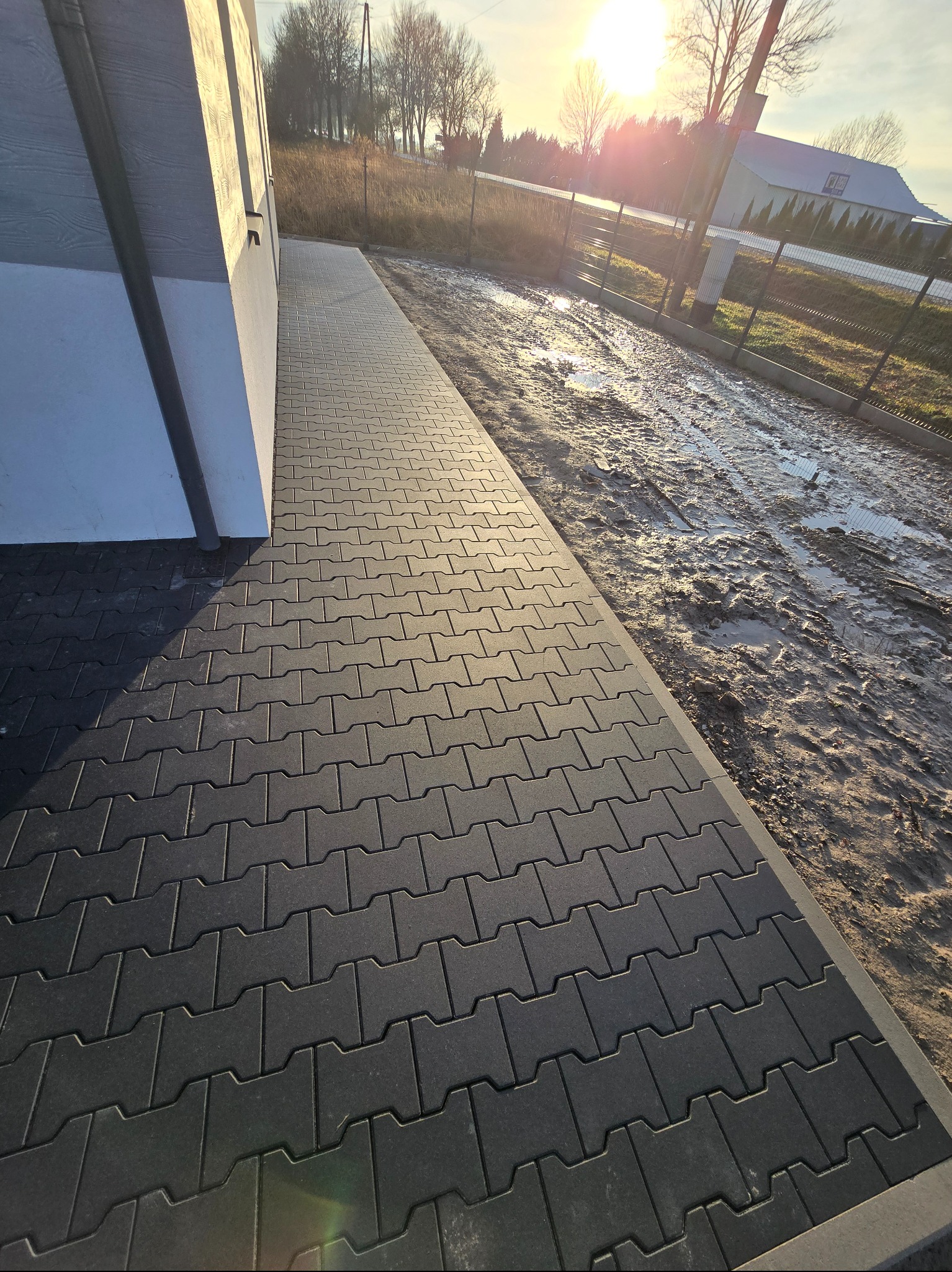 Ułożona, szara kostka brukowa przed budynkiem, z widocznym fragmentem błotnistego terenu obok. Świecące słońce w tle.