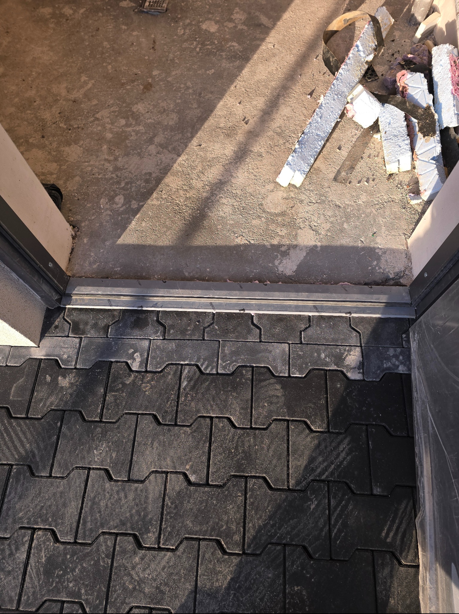 Ułożona, ciemnoszara kostka brukowa przy progu drzwi wejściowych. Widoczne pozostałości prac budowlanych na betonowym podłożu wewnątrz budynku.