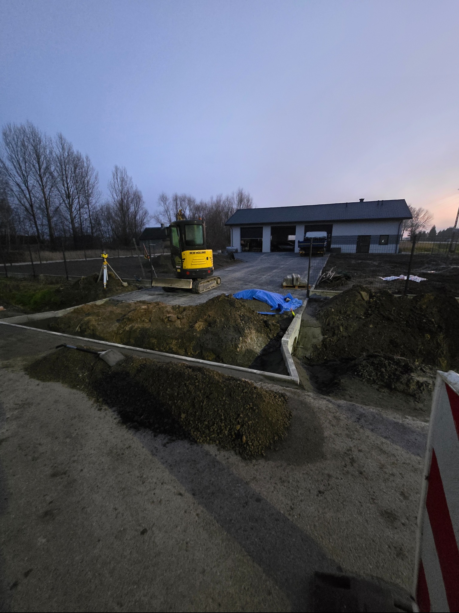 Żółta mini koparka New Holland na placu budowy, obok geodezyjny niwelator laserowy. Widoczne przygotowane fundamenty i hałdy ziemi.