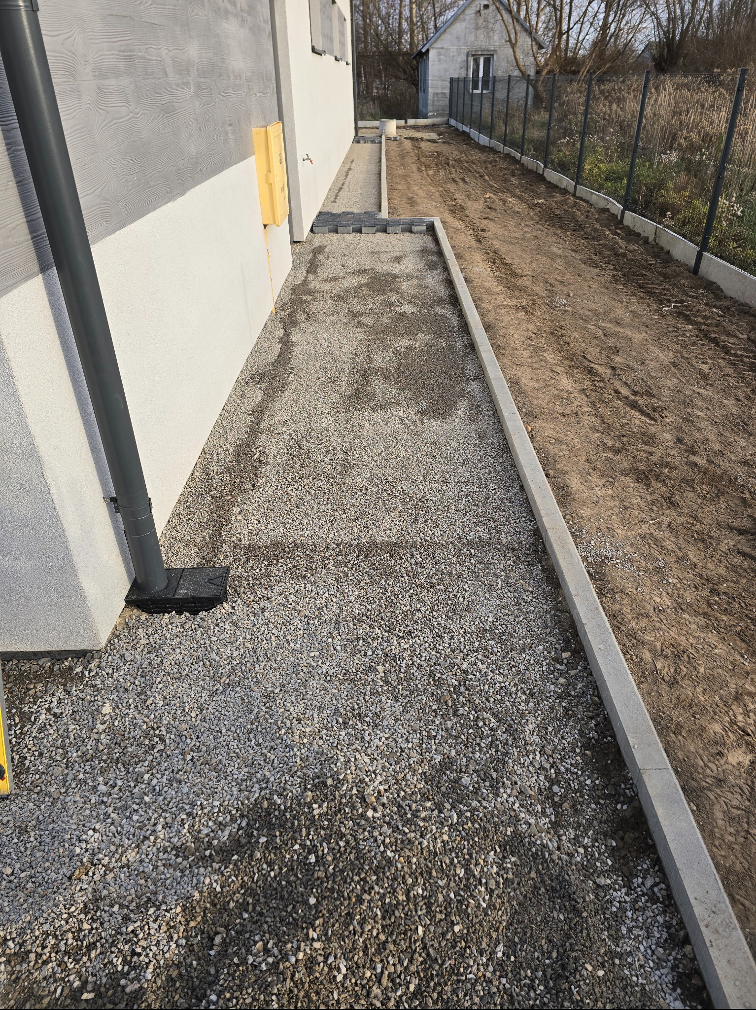 Przygotowanie terenu pod kostkę w Waśniowie: podsypka z kruszywa wzdłuż budynku i obrzeża betonowe. Widoczny fragment rynny spustowej i teren przyszłej drogi.