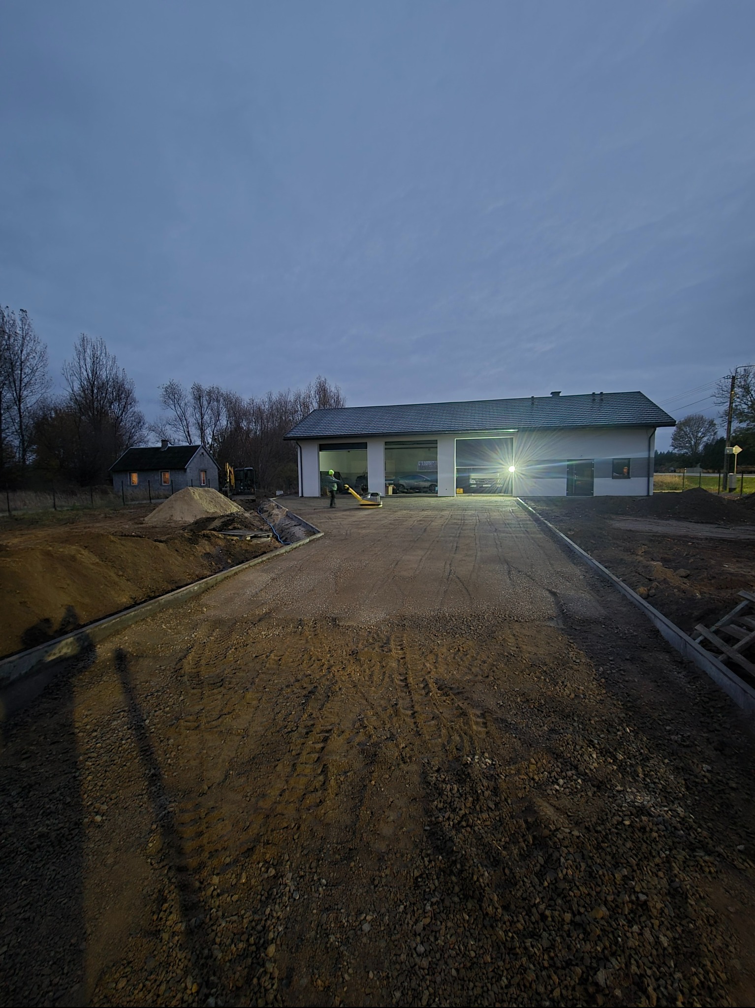 Przygotowany podjazd z utwardzoną nawierzchnią i obrzeżami, w tle budynek z garażem i mały domek, oświetlony teren budowy o zmierzchu.