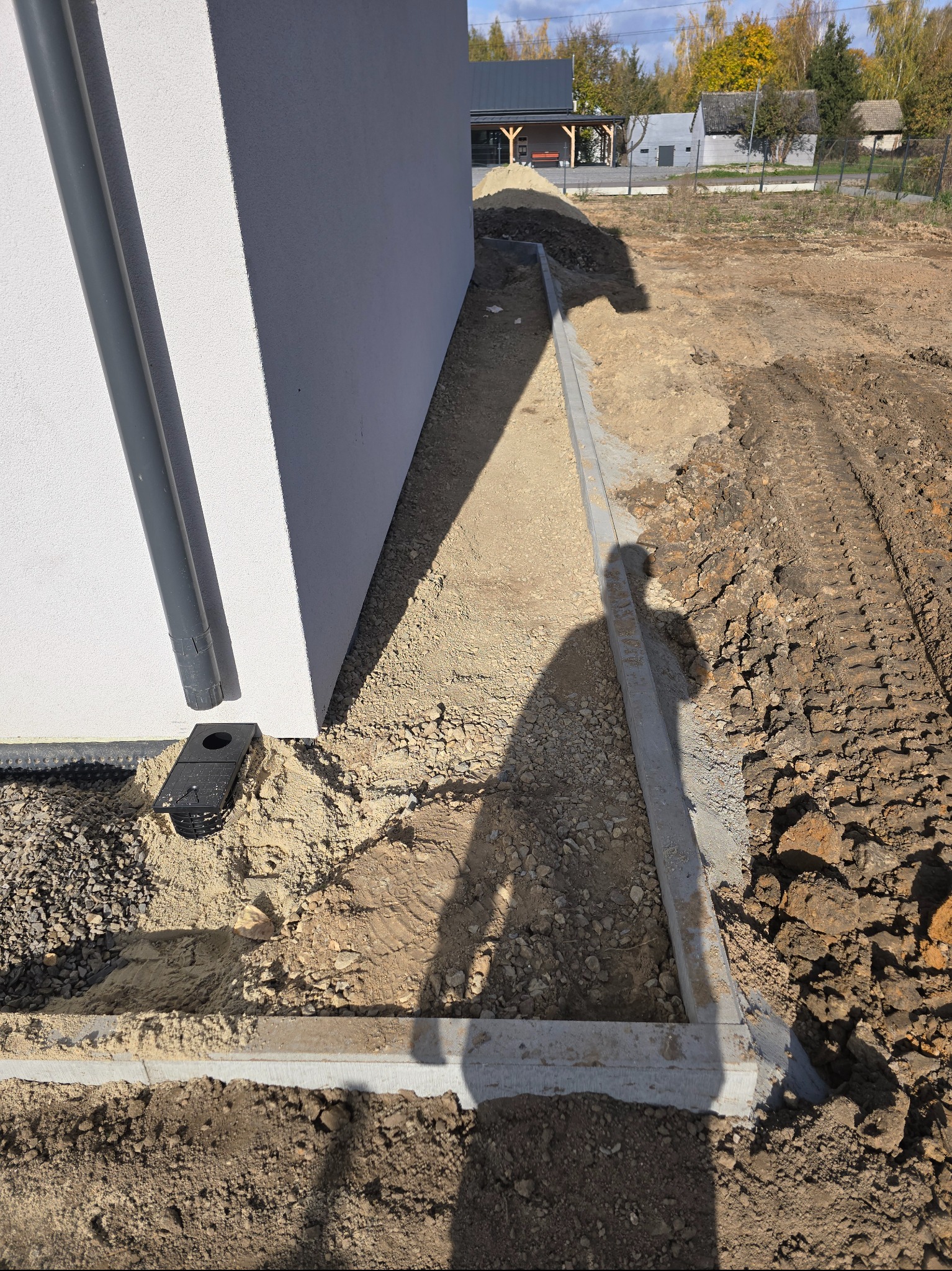 Przygotowanie podłoża pod kostkę brukową przy nowym budynku. Widoczne krawężniki, rury spustowe i częściowo wyrównany teren z shadow.