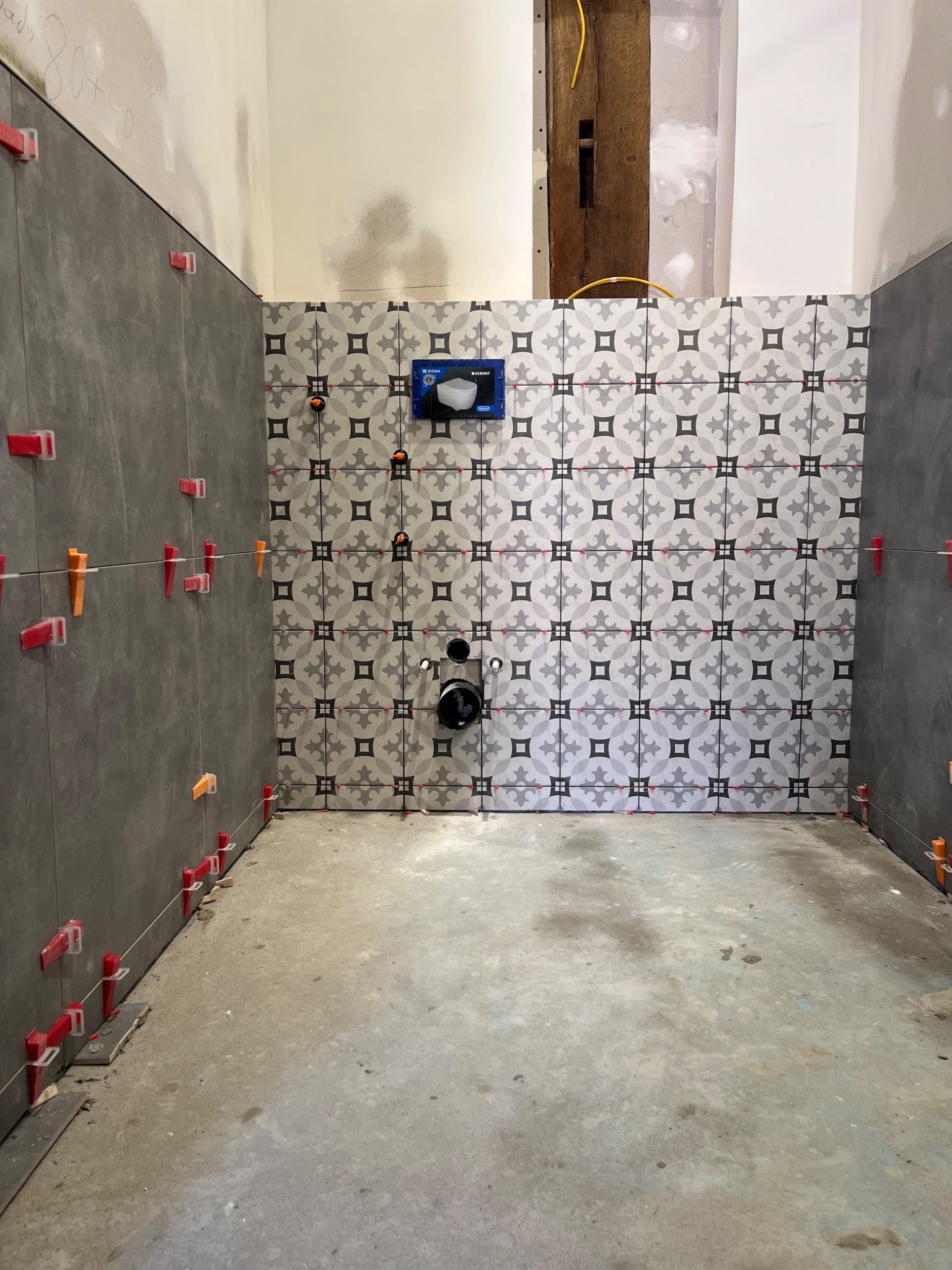 Wnętrze łazienki w trakcie remontu: częściowo ułożone szare płytki na ścianach z systemem poziomowania, dekoracyjne płytki z wzorem na ścianie naprzeciwko, widoczne elementy instalacji sanitarnej.