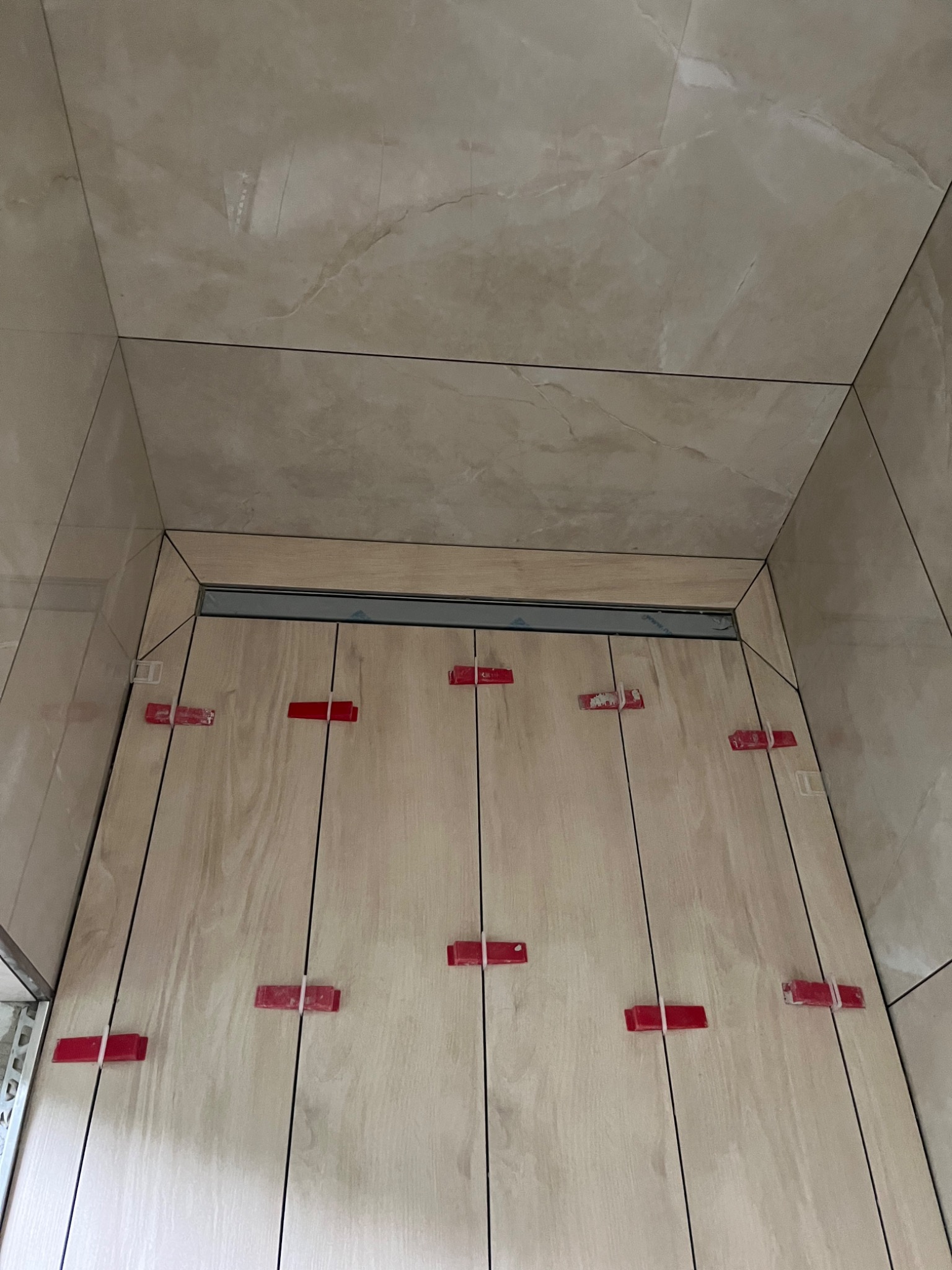 Świeżo ułożone, pionowe płytki imitujące drewno na podłodze prysznica, z widocznymi czerwonymi klipsami poziomującymi i odpływem liniowym w tle.