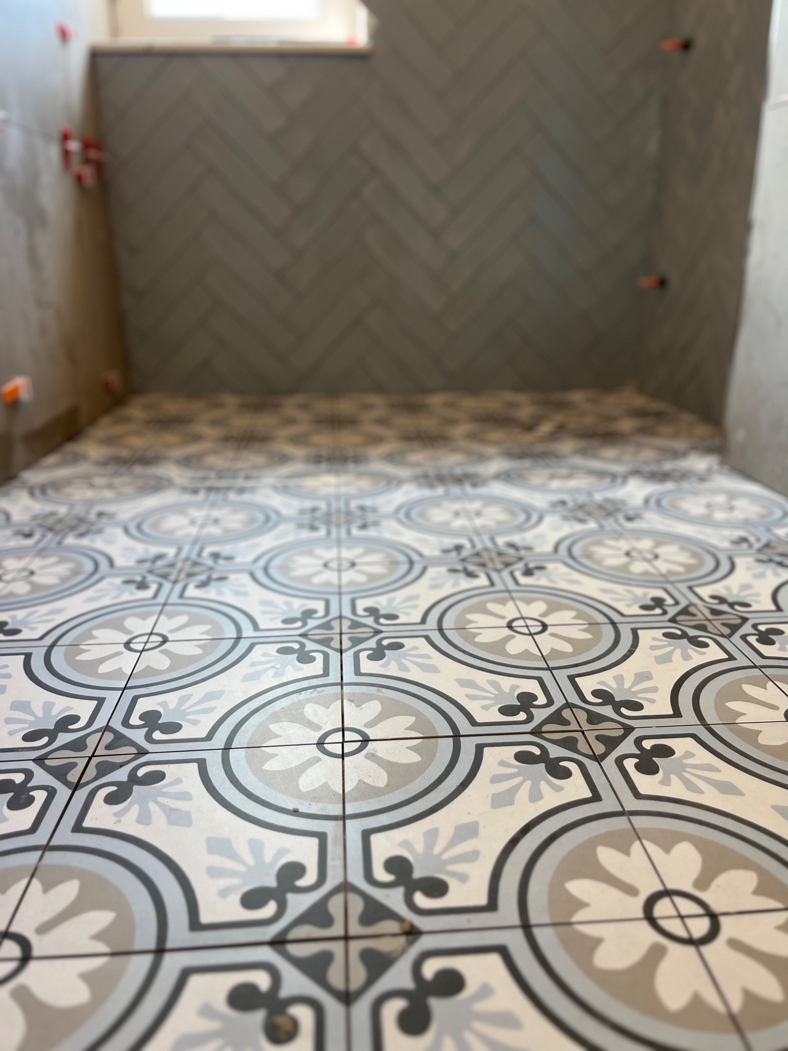 Układanie wzorzystych płytek podłogowych w łazience, z widocznymi krzyżykami dystansowymi, oraz fragmentem ściany wyłożonej szarą cegiełką w jodełkę.