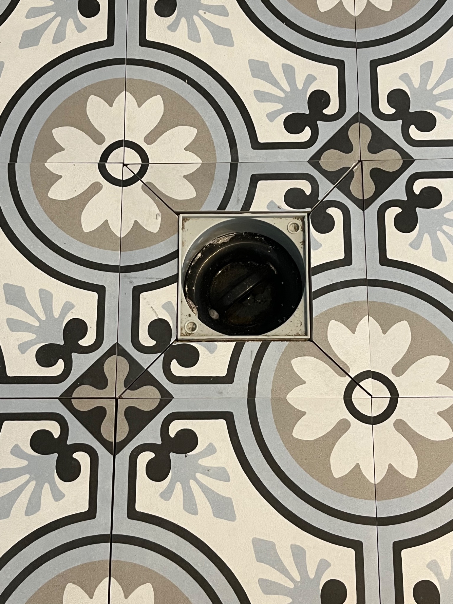 Wzór podłogi z płytek ceramicznych w odcieniach szarości i beżu z centralnie umieszczonym odpływem podłogowym.