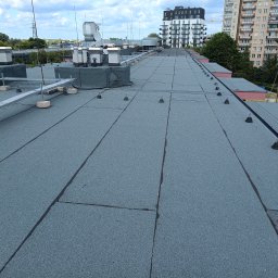 PrimeDach Łukasz Żukowski - Płaski dach pokryty papą termozgrzewalną z elementami instalacji odgromowej i wentylacyjnej na tle zabudowy miejskiej w słoneczny dzień.