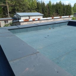 PrimeDach Łukasz Żukowski - Niebieski dach pokryty papą termozgrzewalną z szarymi obróbkami blacharskimi. Widok na budynki i zieleń w tle, słoneczny dzień.