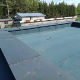 Niebieski dach pokryty papą termozgrzewalną z szarymi obróbkami blacharskimi. Widok na budynki i zieleń w tle, słoneczny dzień.