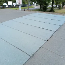 Płaski dach pokryty szarymi panelami bitumicznymi, widoczne łączenia. Na krawędzi dachu widoczne szare płyty. W tle drzewa i tablice informacyjne.