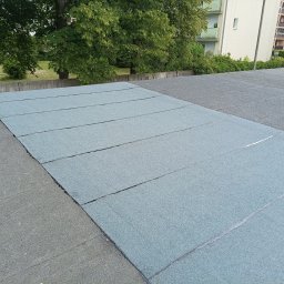 PrimeDach Łukasz Żukowski - Płaski dach pokryty nową, szarozieloną papą bitumiczną. Widoczne połączenia między pasami papy. W tle drzewa i budynki mieszkalne.