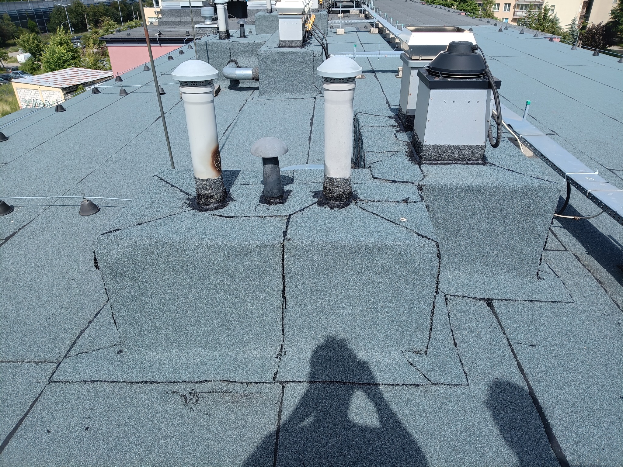 Płaski dach pokryty papą z kominami wentylacyjnymi i elementami instalacji. Widoczny cień osoby robiącej zdjęcie. Dach w odcieniach szarości i błękitu.