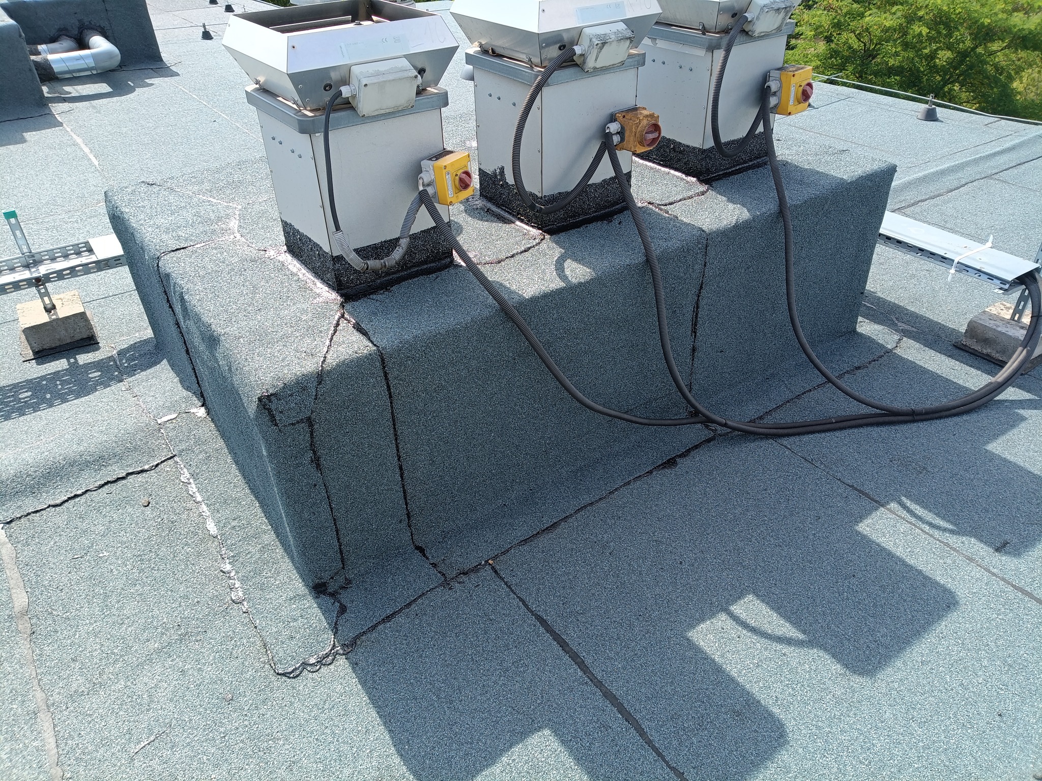 Płaski dach pokryty papą z trzema kominami wentylacyjnymi i instalacją elektryczną. Widoczne pęknięcia na papie, montaż instalacji w toku.