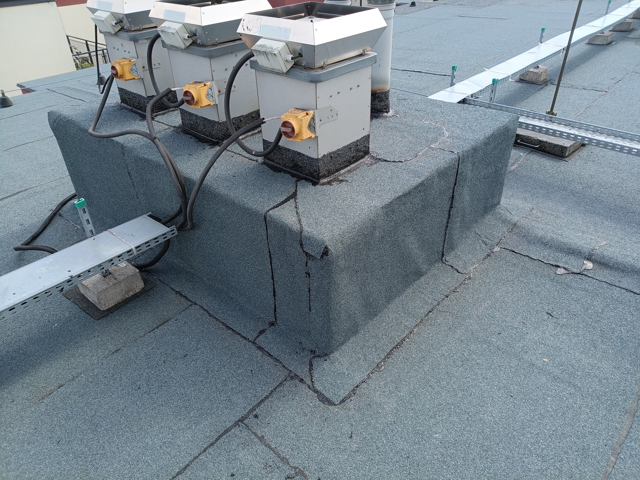 Płaski dach pokryty papą z zabudowanymi elementami instalacji wentylacyjnej i metalowymi korytkami kablowymi. Widoczne uszczelnienia i łączenia papy.