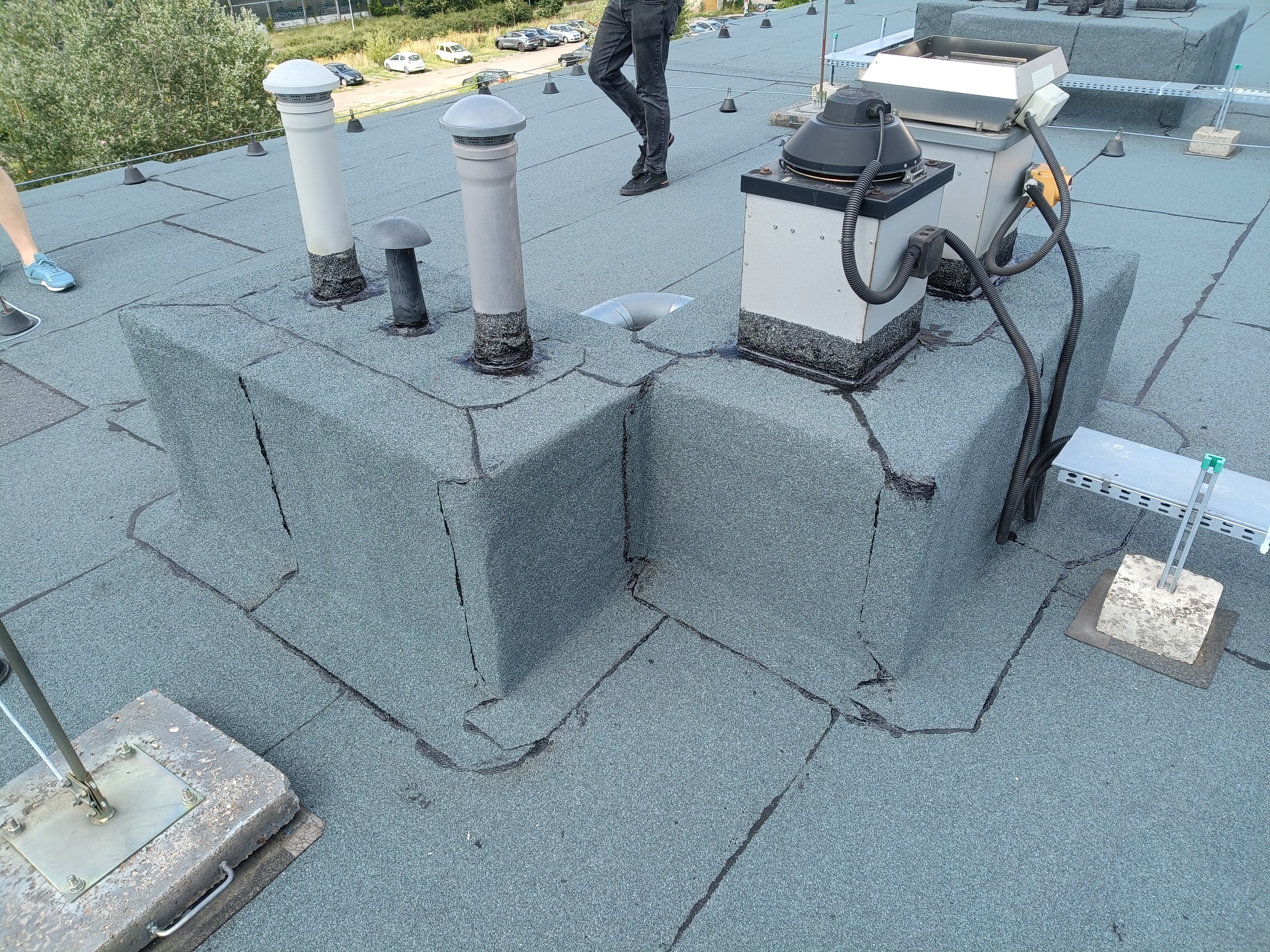 Szczegółowe ujęcie płaskiego dachu pokrytego papą z kominami, wentylacją i elementami instalacji, z widocznymi łączeniami i teksturą powierzchni.