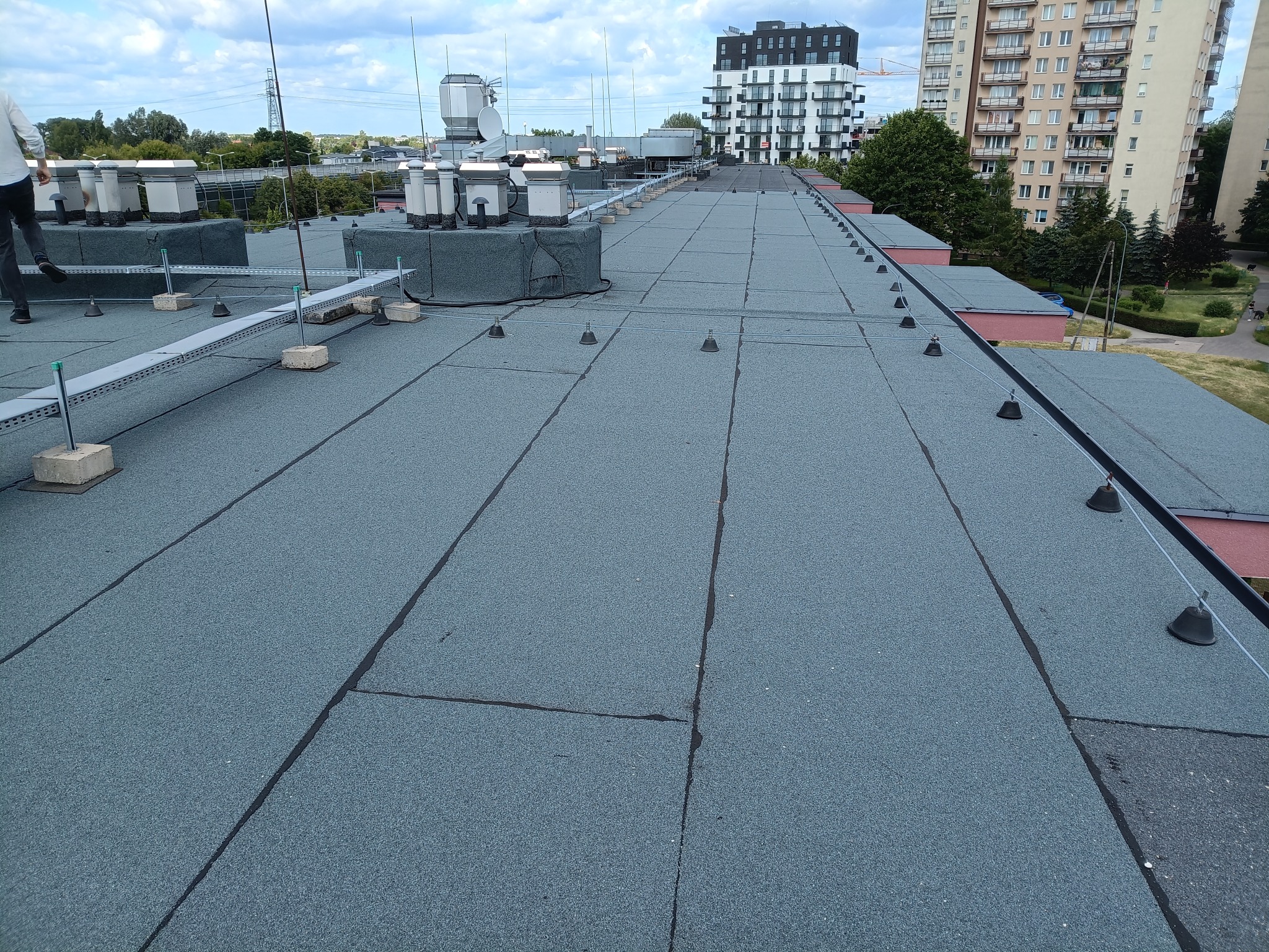 Płaski dach pokryty papą termozgrzewalną z elementami instalacji odgromowej i wentylacyjnej na tle zabudowy miejskiej w słoneczny dzień.