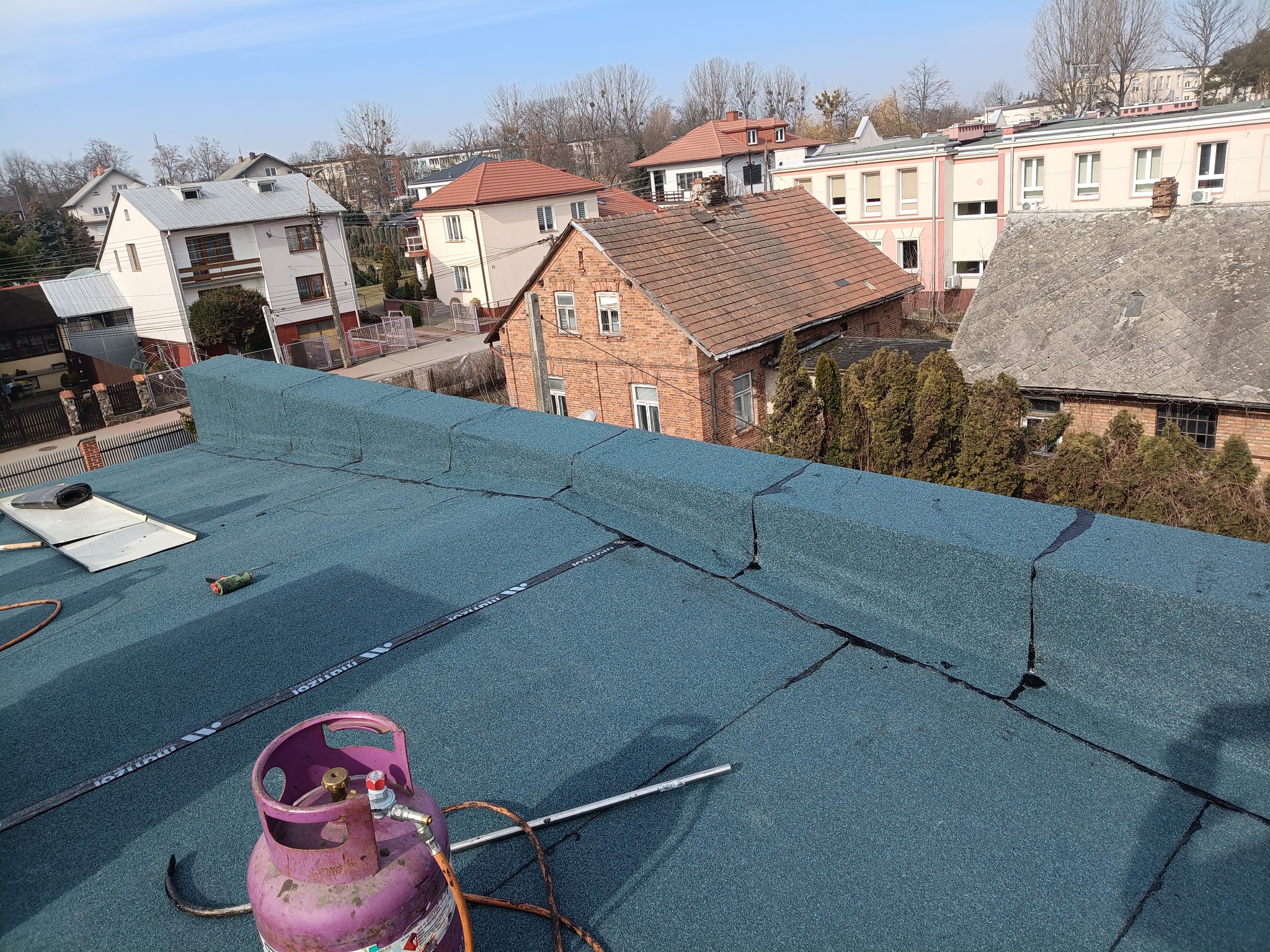 Niebieski dach pokryty papą termozgrzewalną z widocznym sprzętem dekarskim i zabudową komina, w tle zabudowa miejska w słoneczny dzień.