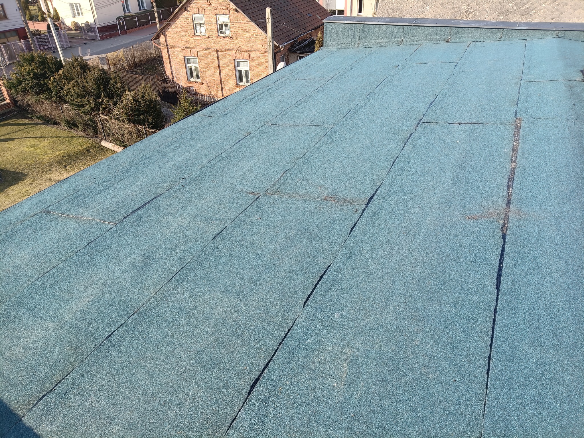 Niebieski dach płaski pokryty papą termozgrzewalną, widoczne łączenia i faktura materiału. W tle zabudowa jednorodzinna i zieleń. Słoneczny dzień.