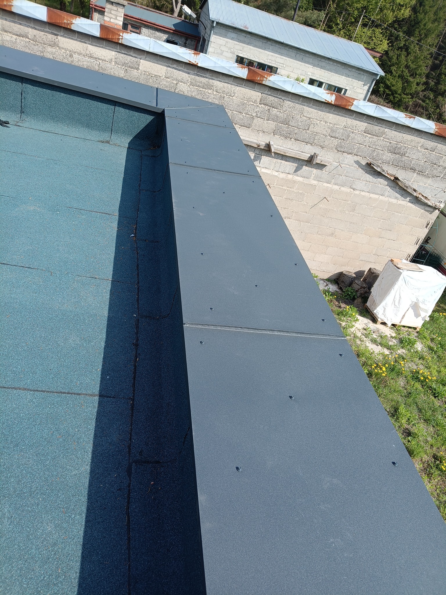 Szczegółowe ujęcie płaskiego dachu pokrytego papą i obróbką blacharską w kolorze grafitowym, z widocznym budynkiem w tle i zielenią w otoczeniu.