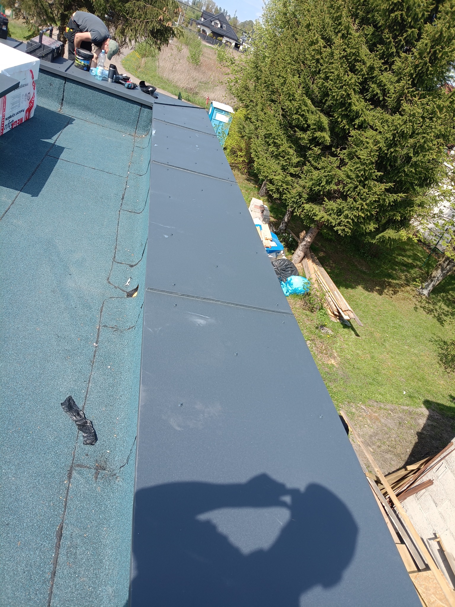 Dekarz pracuje na dachu pokrytym papą, montując obróbki blacharskie. Widoczne narzędzia, materiały i cień pracownika. Ujęcie z góry, w tle zieleń drzew i zabudowania.