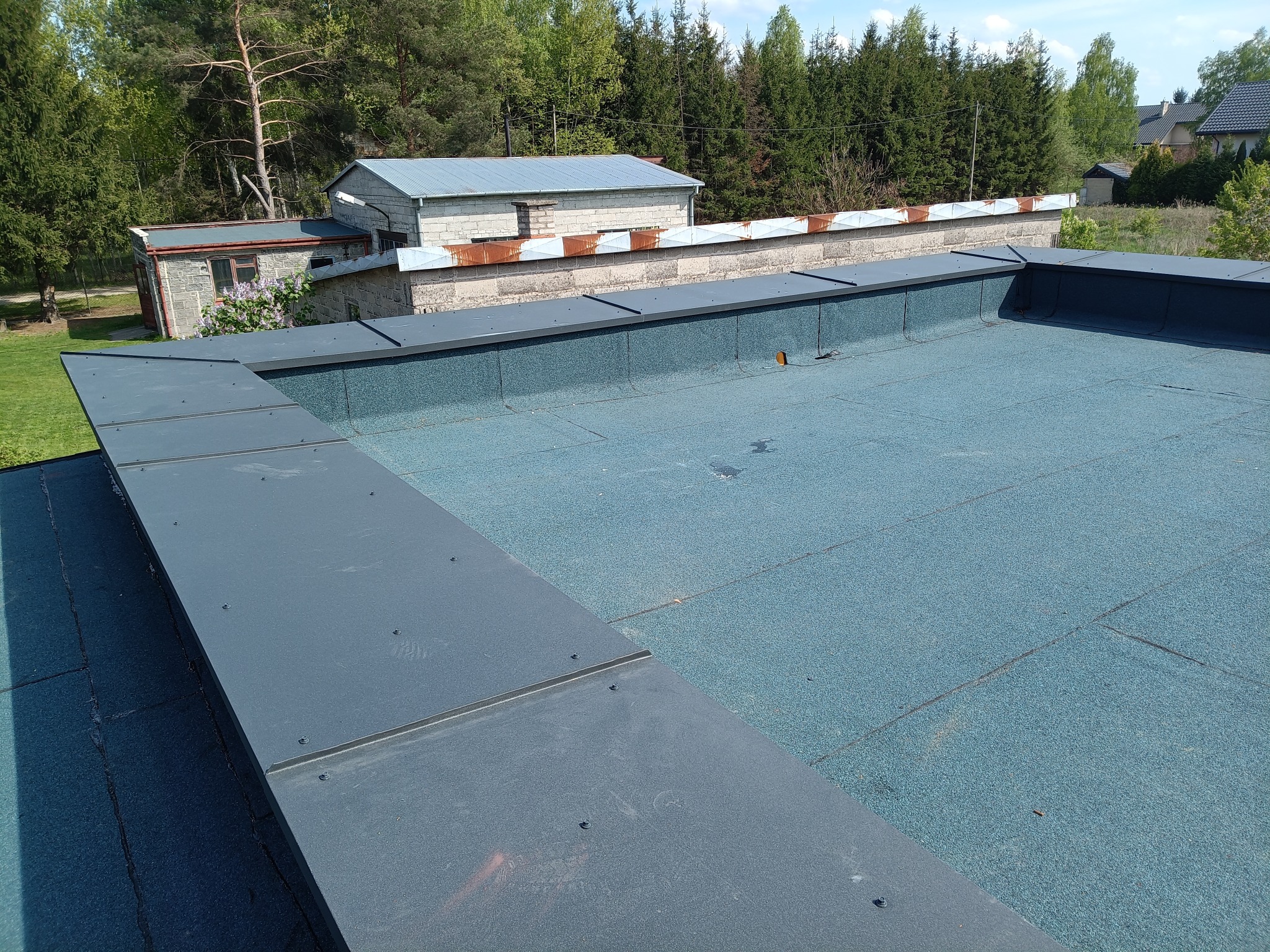 Niebieski dach pokryty papą termozgrzewalną z szarymi obróbkami blacharskimi. Widok na budynki i zieleń w tle, słoneczny dzień.
