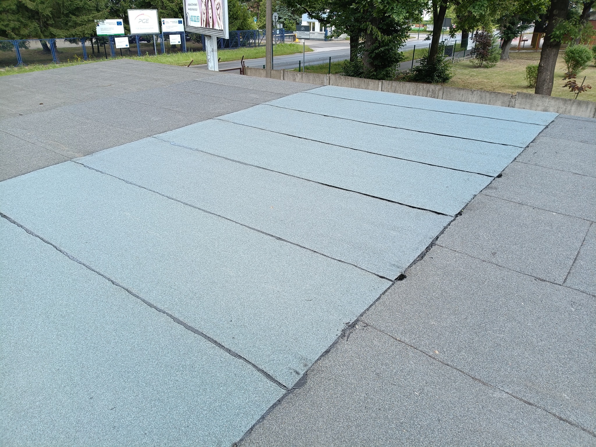 Płaski dach pokryty szarymi panelami bitumicznymi, widoczne łączenia. Na krawędzi dachu widoczne szare płyty. W tle drzewa i tablice informacyjne.