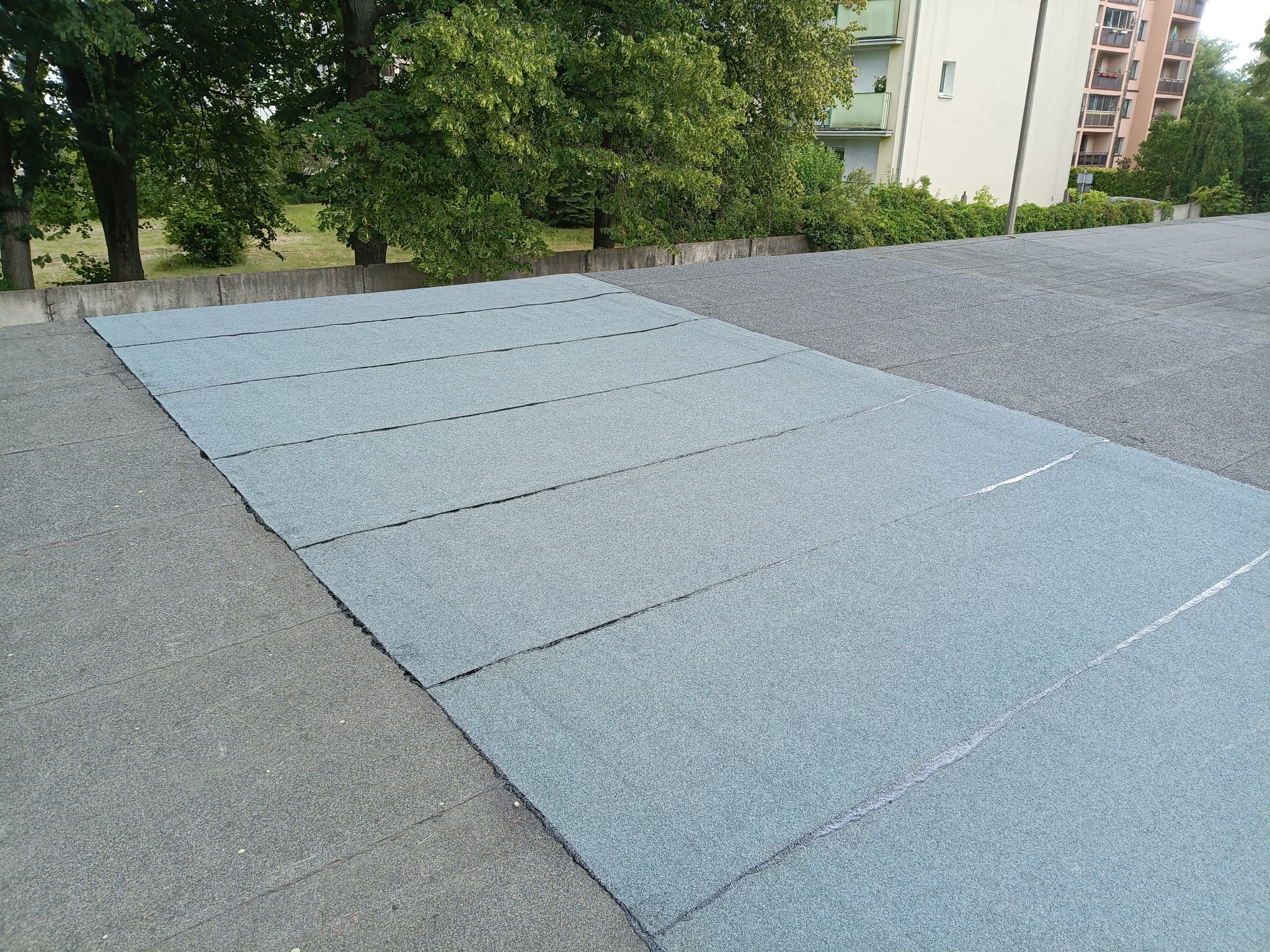 Płaski dach pokryty nową, szarozieloną papą bitumiczną. Widoczne połączenia między pasami papy. W tle drzewa i budynki mieszkalne.