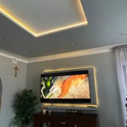 Szary sufit z podwieszaną konstrukcją i oświetleniem LED, telewizor z podświetleniem, drewniana komoda, sztuczna roślina w rogu pokoju, krzyż na ścianie.