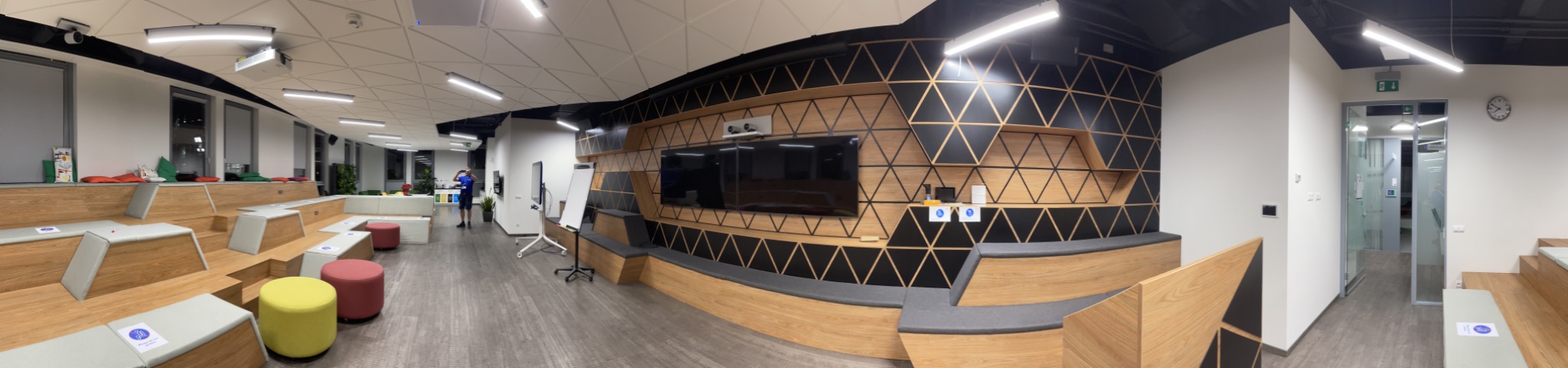 Panoramiczny widok nowocześnie urządzonej przestrzeni biurowej z drewnianymi schodkowymi siedziskami, poduszkami, pufami i ścianą z geometrycznym wzorem trójkątów, oświetlonej lampami sufitowymi.