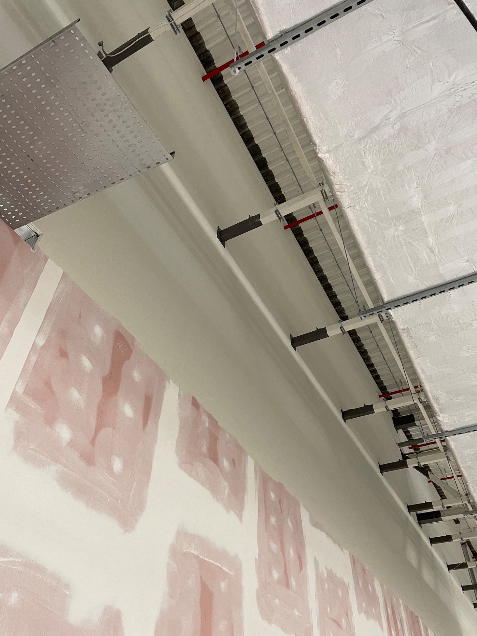 Przygotowana do malowania ściana z płyt kartonowo-gipsowych z widocznymi łączeniami szpachlowanymi na różowo, w tle konstrukcja sufitu podwieszanego z izolacją.