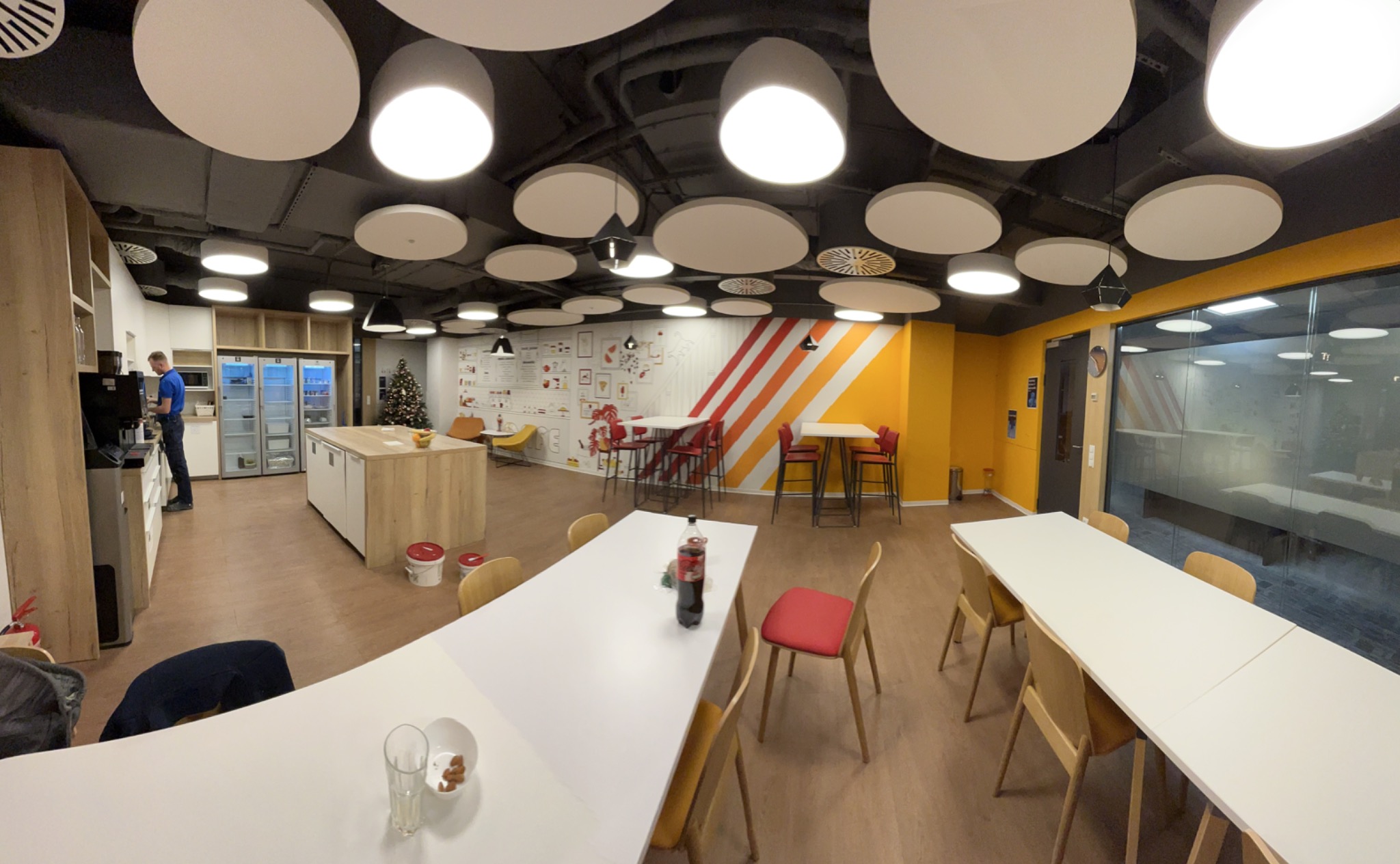 Panoramiczne ujęcie nowoczesnej przestrzeni biurowej z białymi stołami, kolorowymi krzesłami, okrągłymi lampami sufitowymi oraz ścianami z dekoracyjnymi wzorami i pomalowanymi w pasy, widoczny...