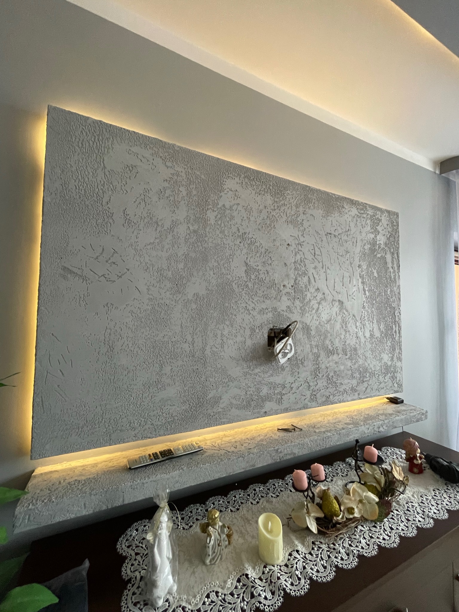 Dekoracyjna ściana TV z oświetleniem LED, fakturą betonu architektonicznego i półką, na której leży pilot; na komodzie ozdobna serweta z figurkami i świecami.