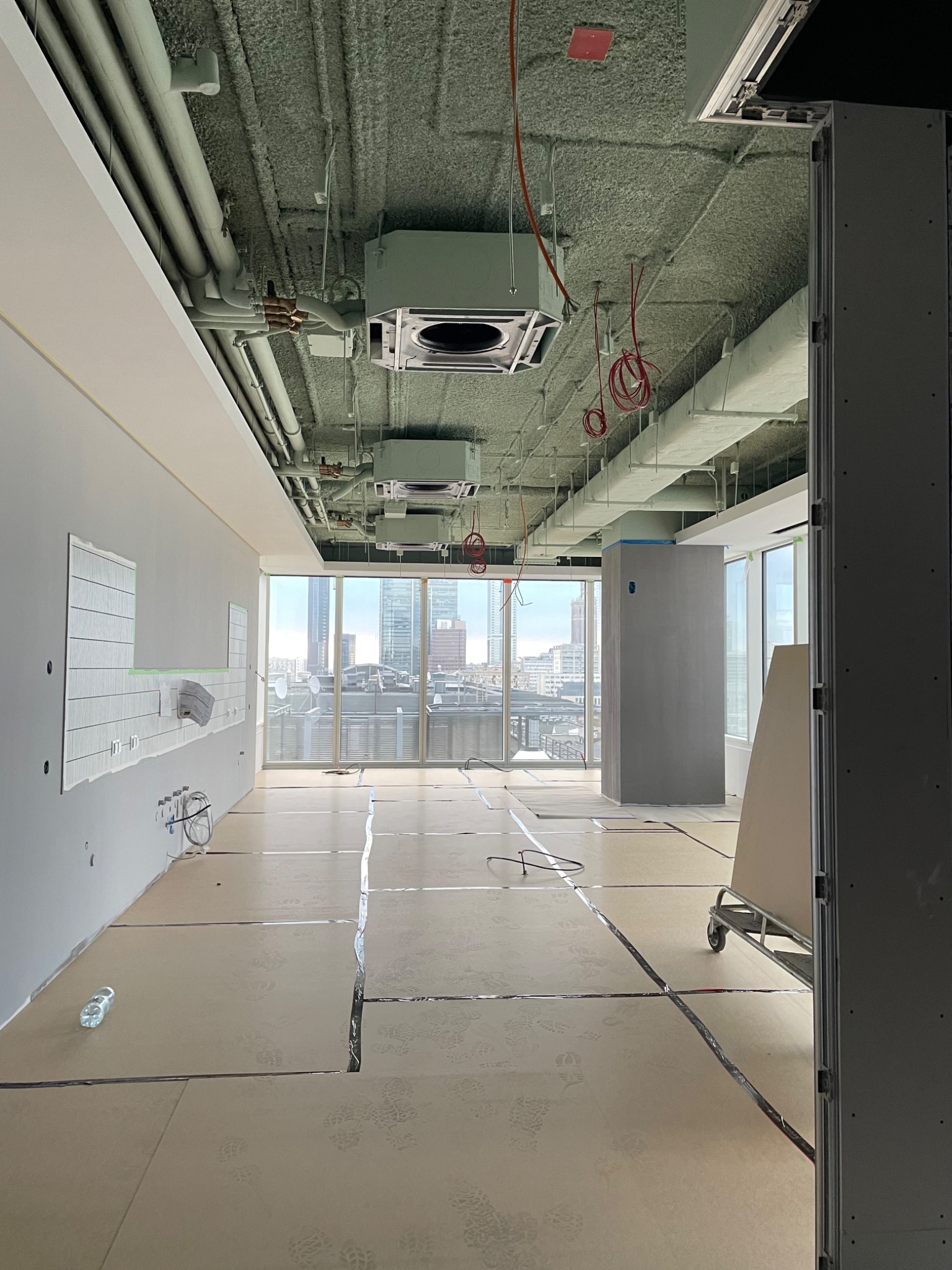 Wnętrze biurowe w trakcie prac wykończeniowych: sufit pokryty natryskową izolacją, przygotowane podłoże pod panele, instalacje pod sufitem, widok na miasto za oknem.